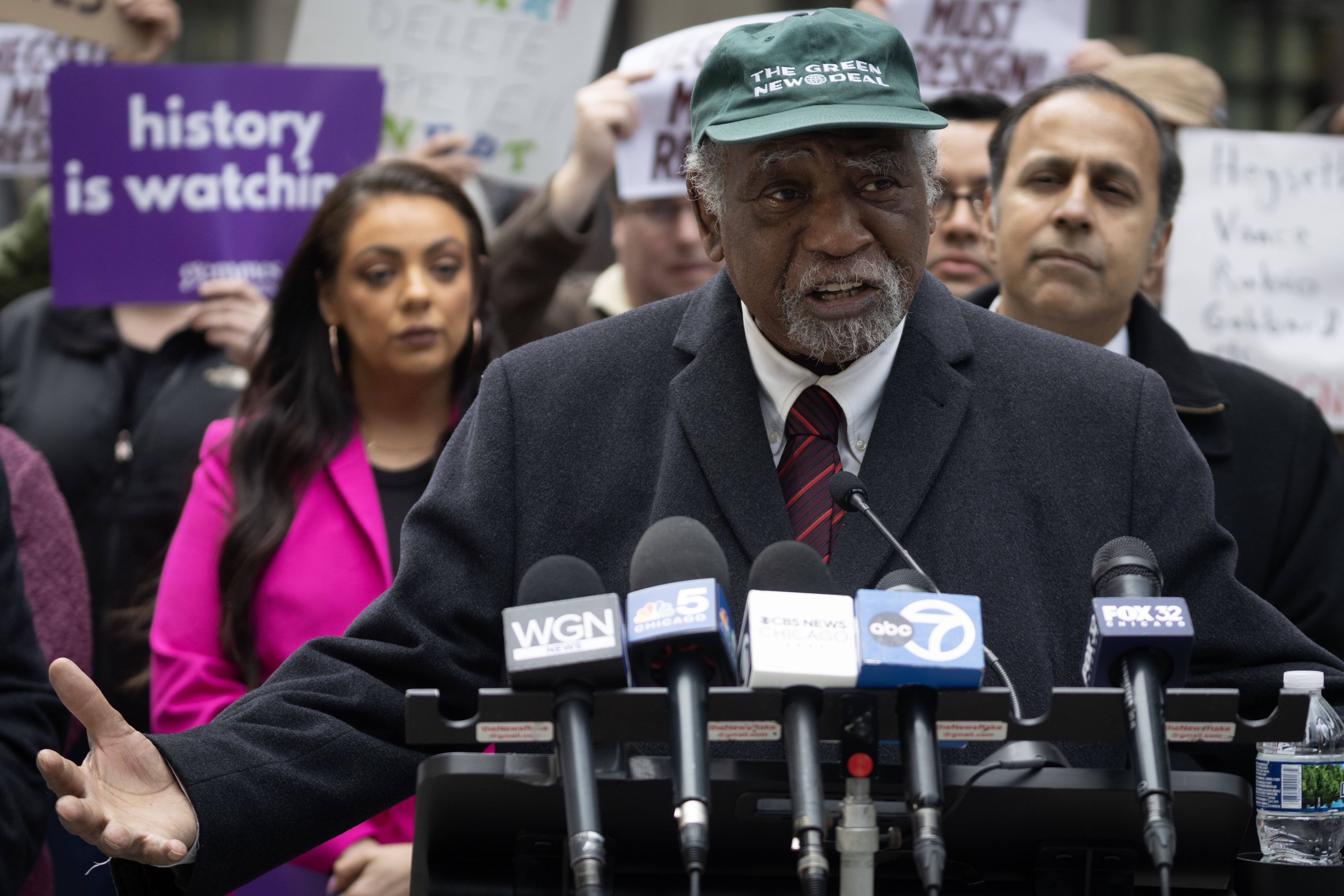 A man with gray hair and beard wearing a dark coat, red striped tie, and green cap that reads "THE GREEN NEW DEAL" speaks at a podium with multiple media microphones. People hold protest signs behind him, including one purple sign that says "history is watching."