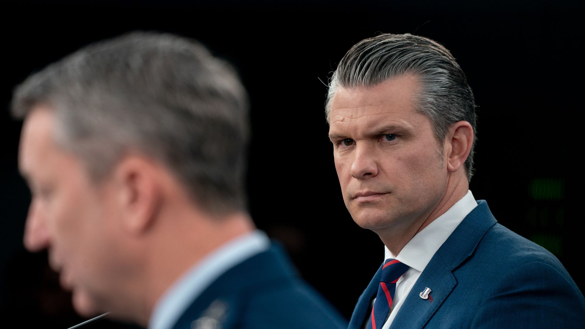 Pete Hegseth — wearing a dark suit, a white collared shirt and a striped blue and red tie — looks on while Dan Caine speaks at a press conference.