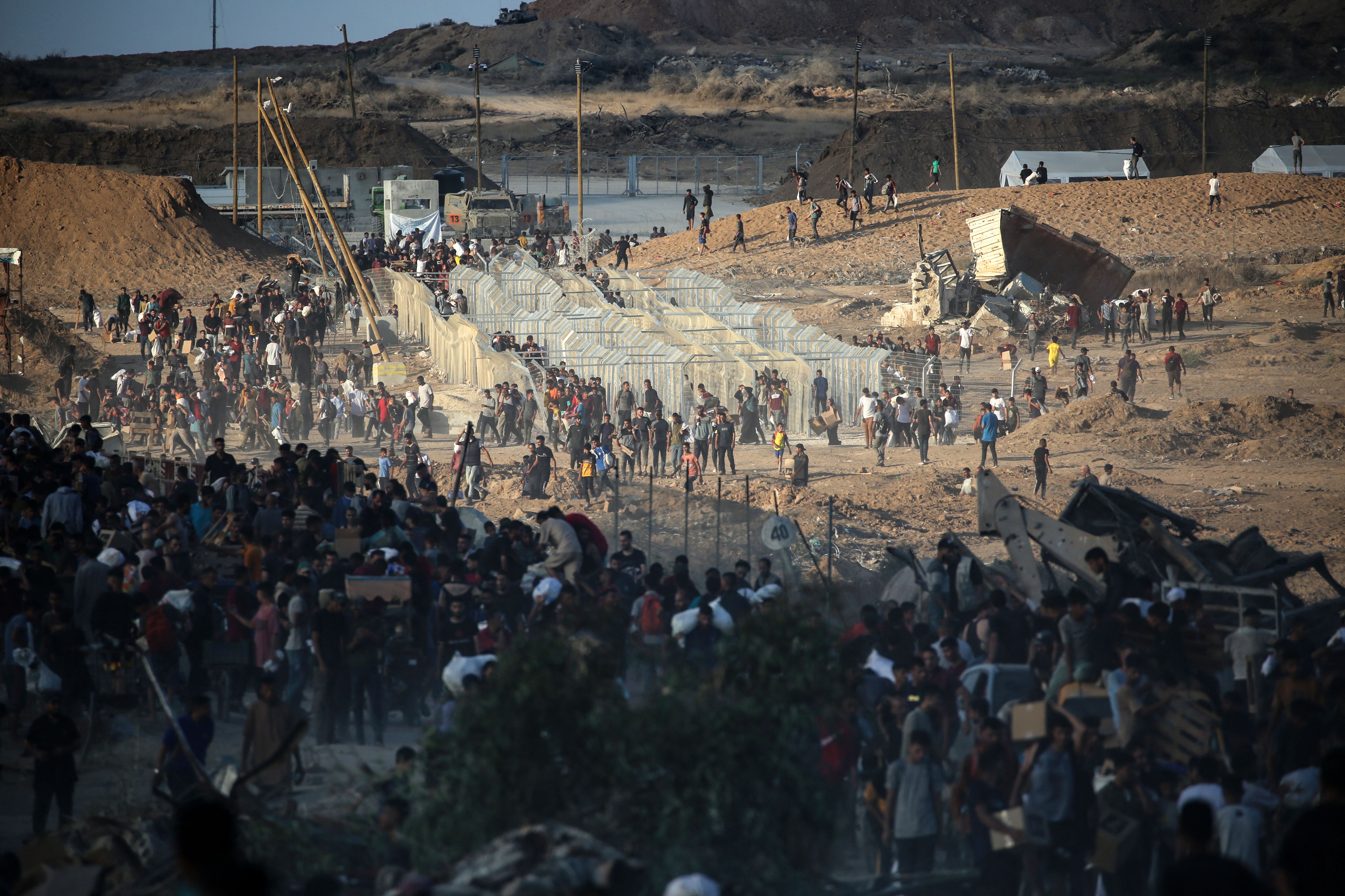 Large crowds of people walking near and around a fenced-off checkpoint in a dusty, hilly area with some tents, vehicles, and construction equipment visible.
