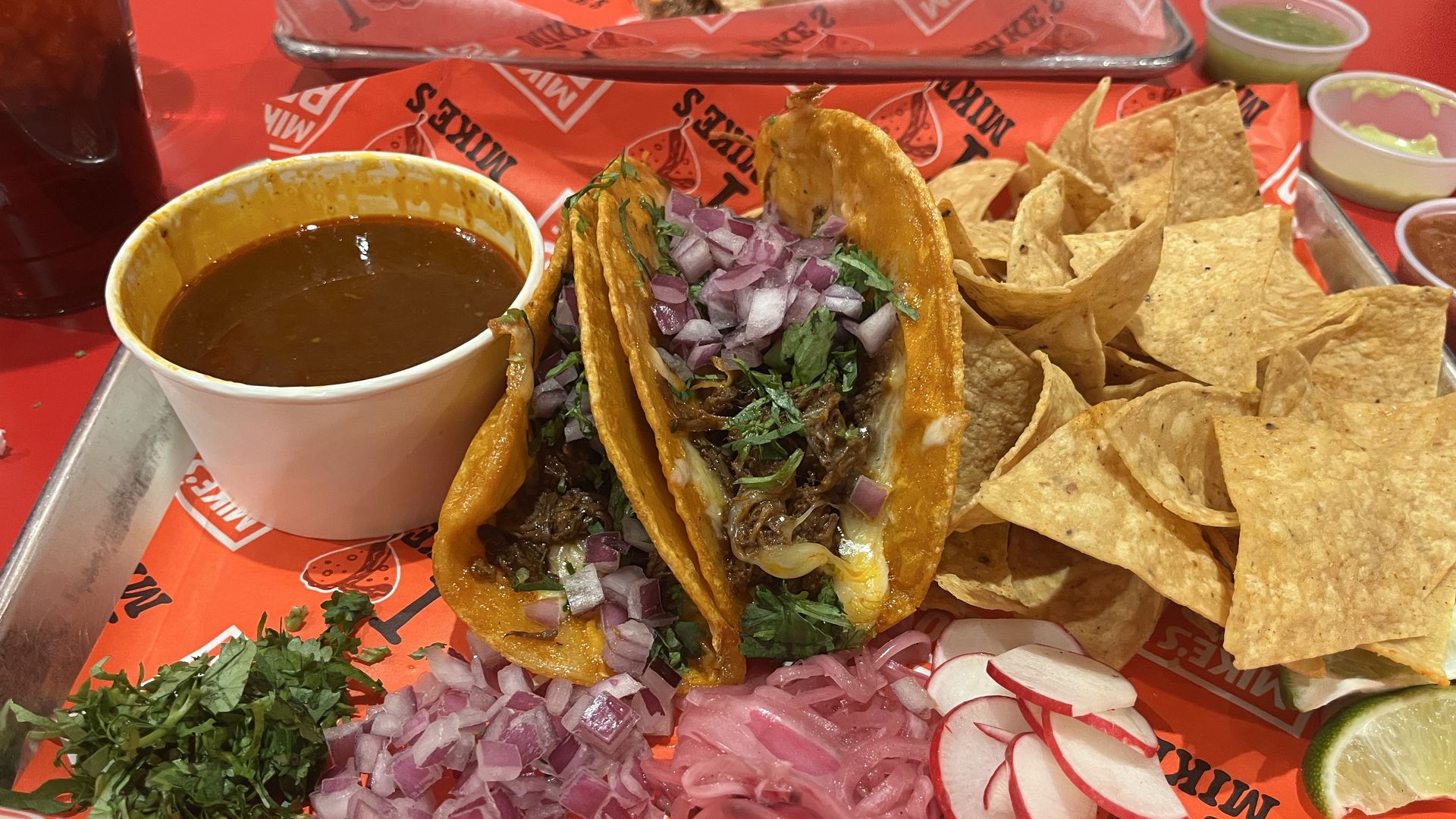 Tray with two beef birria tacos filled with onions, cilantro, and cheese, surrounded by chopped red onions, radish slices, lime wedge, chopped herbs, and a pile of tortilla chips with a cup of brown sauce.
