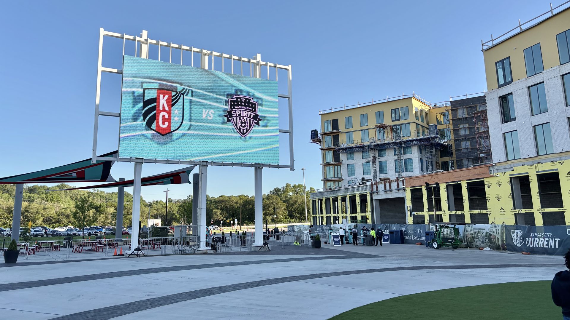 Outdoor plaza with a large LED screen displaying KC vs Spirit logos against a blue sky. Right side shows a building under construction with scaffolding; barriers and people near the entrance.