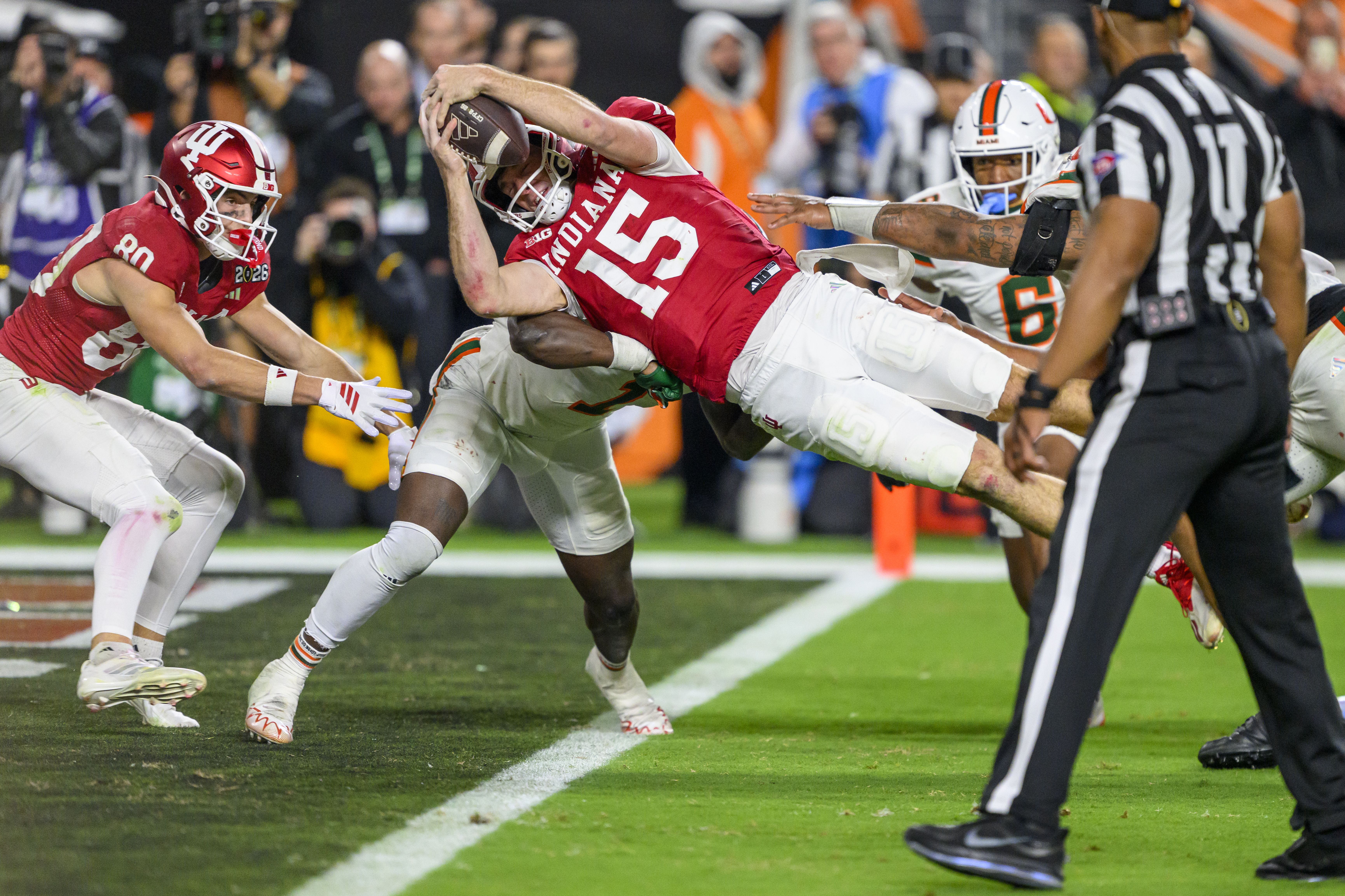 Football player in red Indiana uniform with number 15 dives to catch ball near sideline while white-uniformed Miami players tackle him, with referee watching closely on green field.