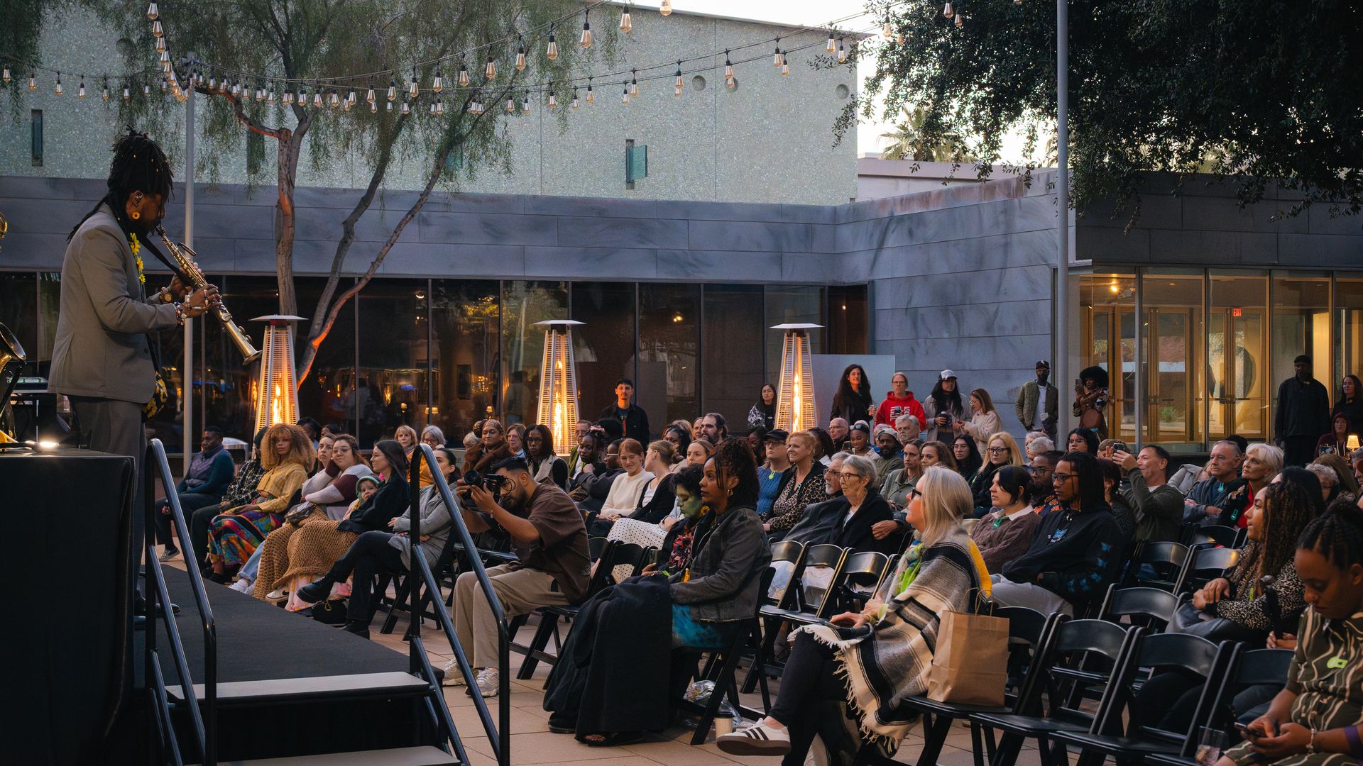 Outdoor concert with a saxophonist in a gray suit playing on stage to a seated audience beneath string bulb lights and warm heaters, with trees and a modern building in the background.