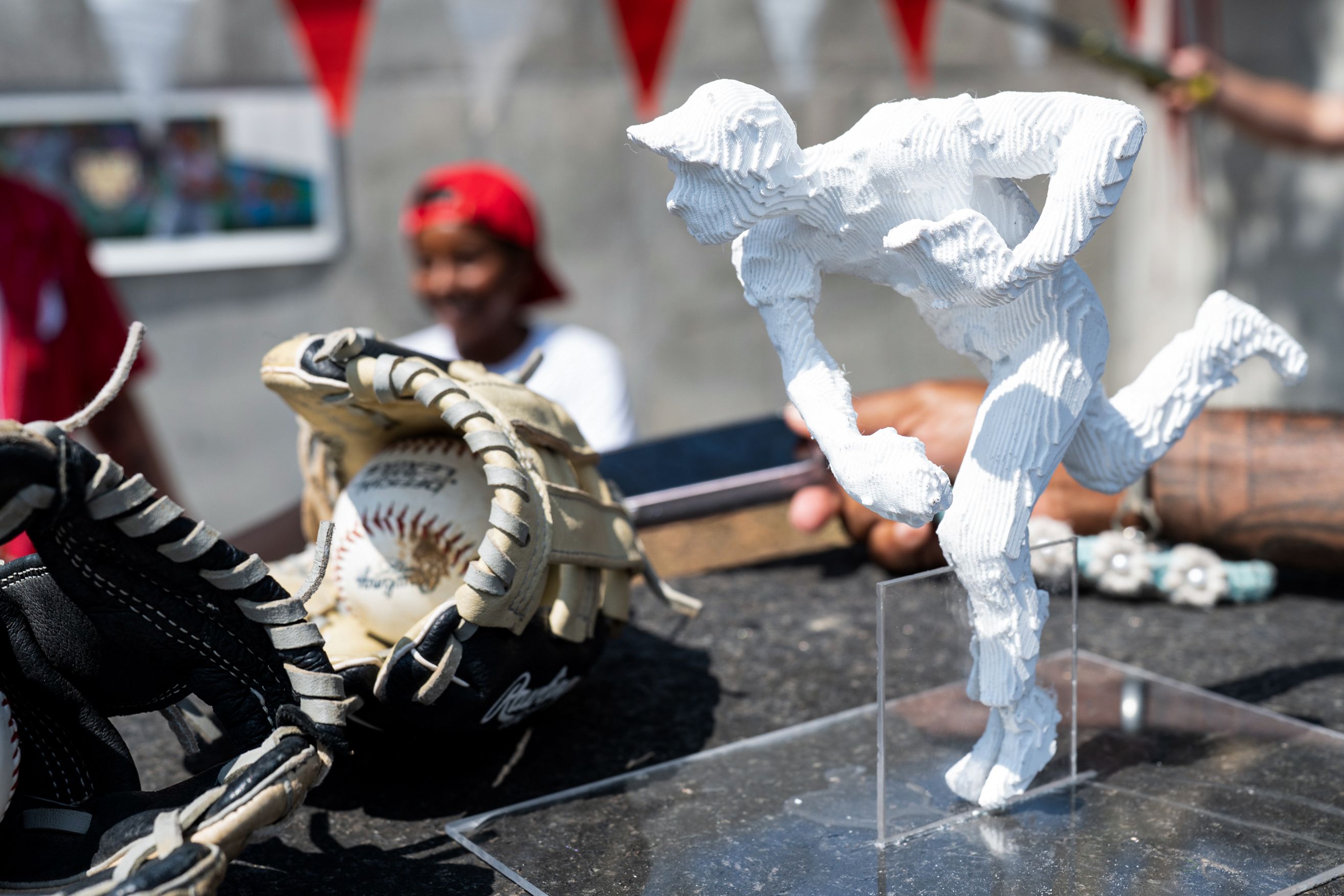 White textured fabric sculpture in the foreground, with a worn baseball glove and baseball nearby; a blurred figure wearing a red cap and red bunting in the background.