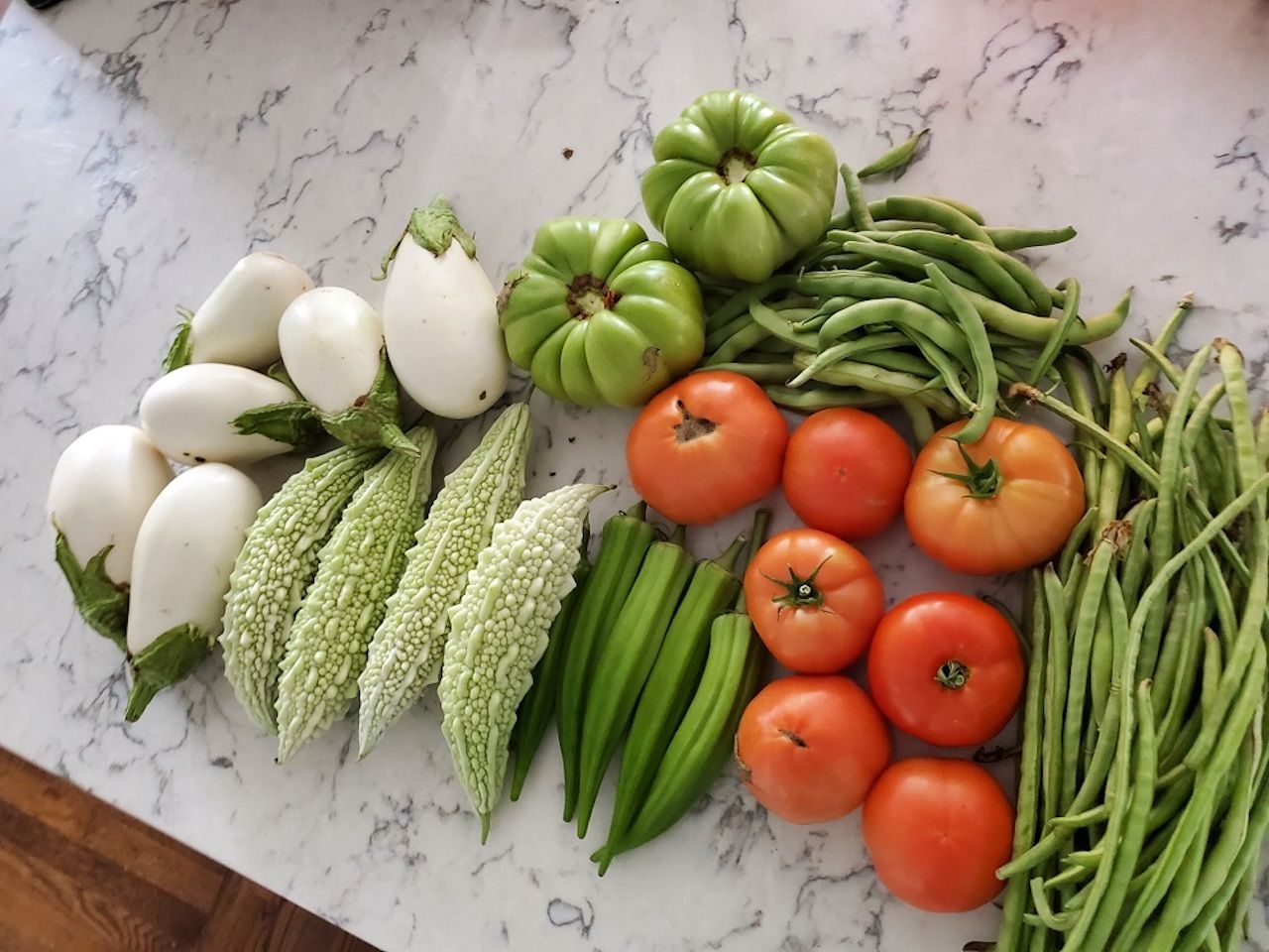 Assorted fresh vegetables on a marble counter: white eggplants, pale green bitter melons, green beans, okra, ribbed green tomatoes, and orange tomatoes.