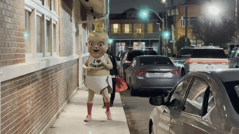 Image shows the King Cake Baby mascot lurking and walking on a sidewalk with cars passing by.