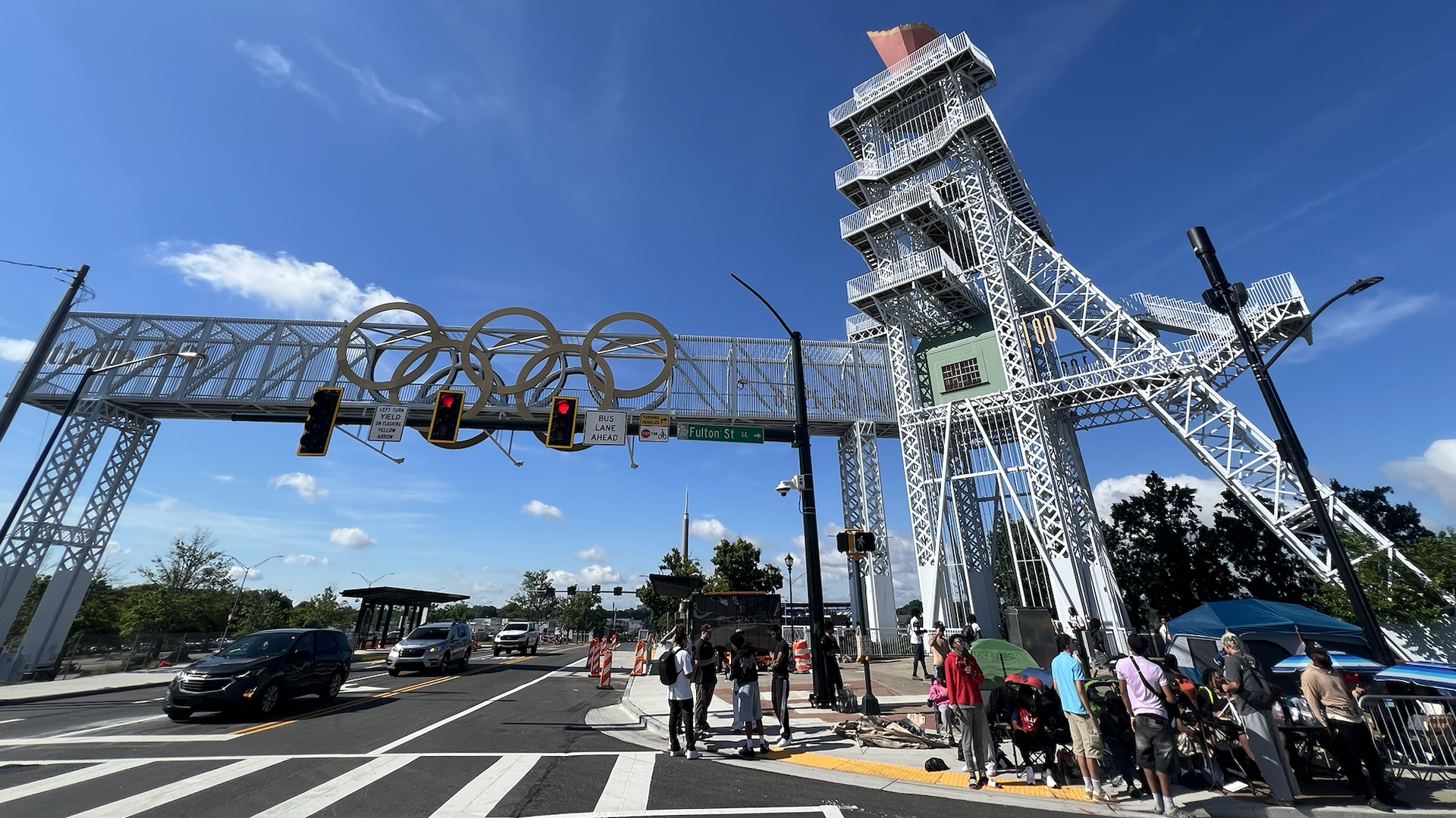 Clear blue sky over a white steel observation tower and elevated pedestrian walkway decorated with large Olympic rings. Cars travel below as people stand at the crosswalk.