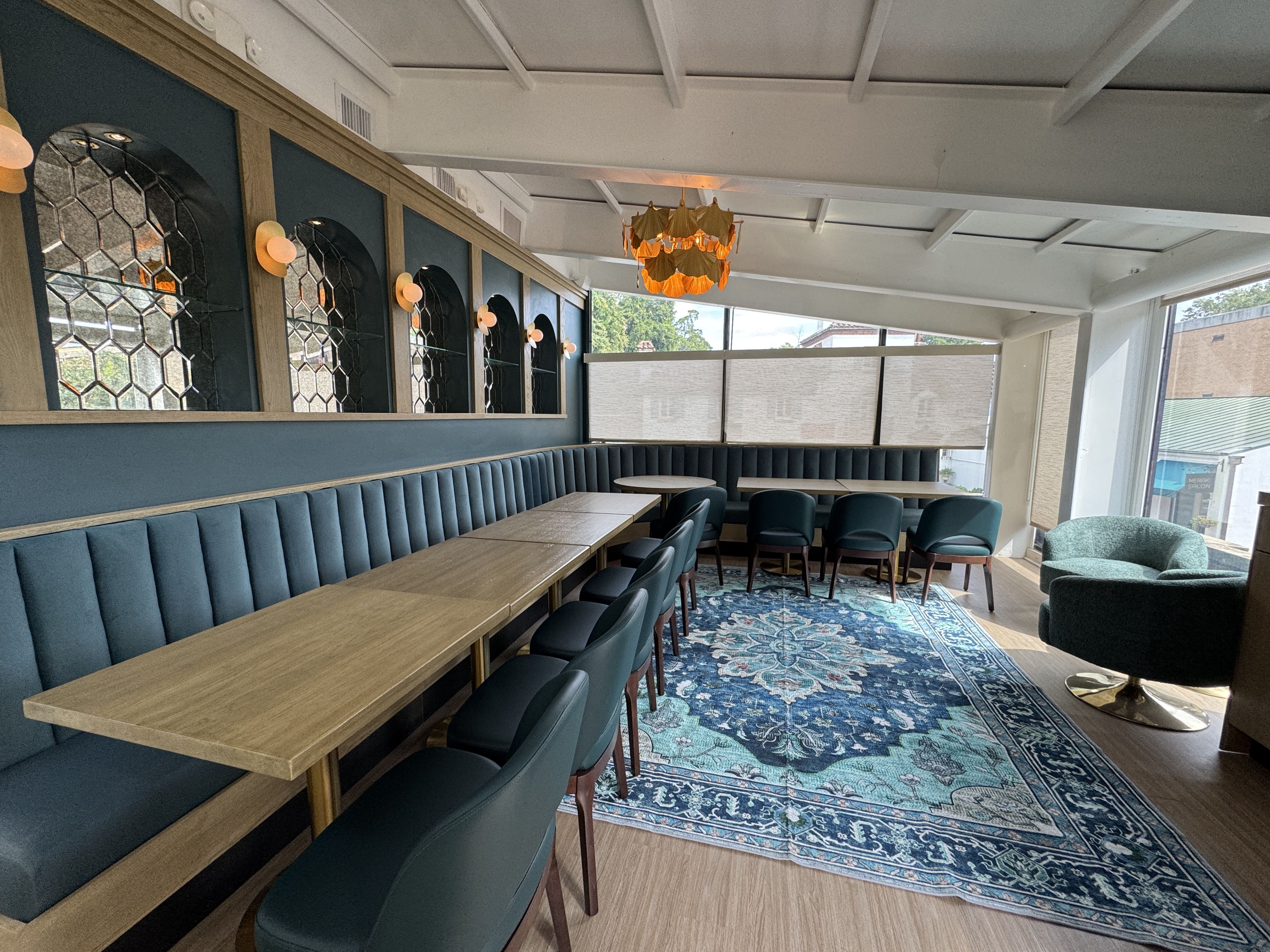 Modern seating area with teal cushioned bench and chairs, light wood tables, blue patterned rug, honeycomb mirrors, soft round wall lights, and a hanging orange light fixture in a bright room.