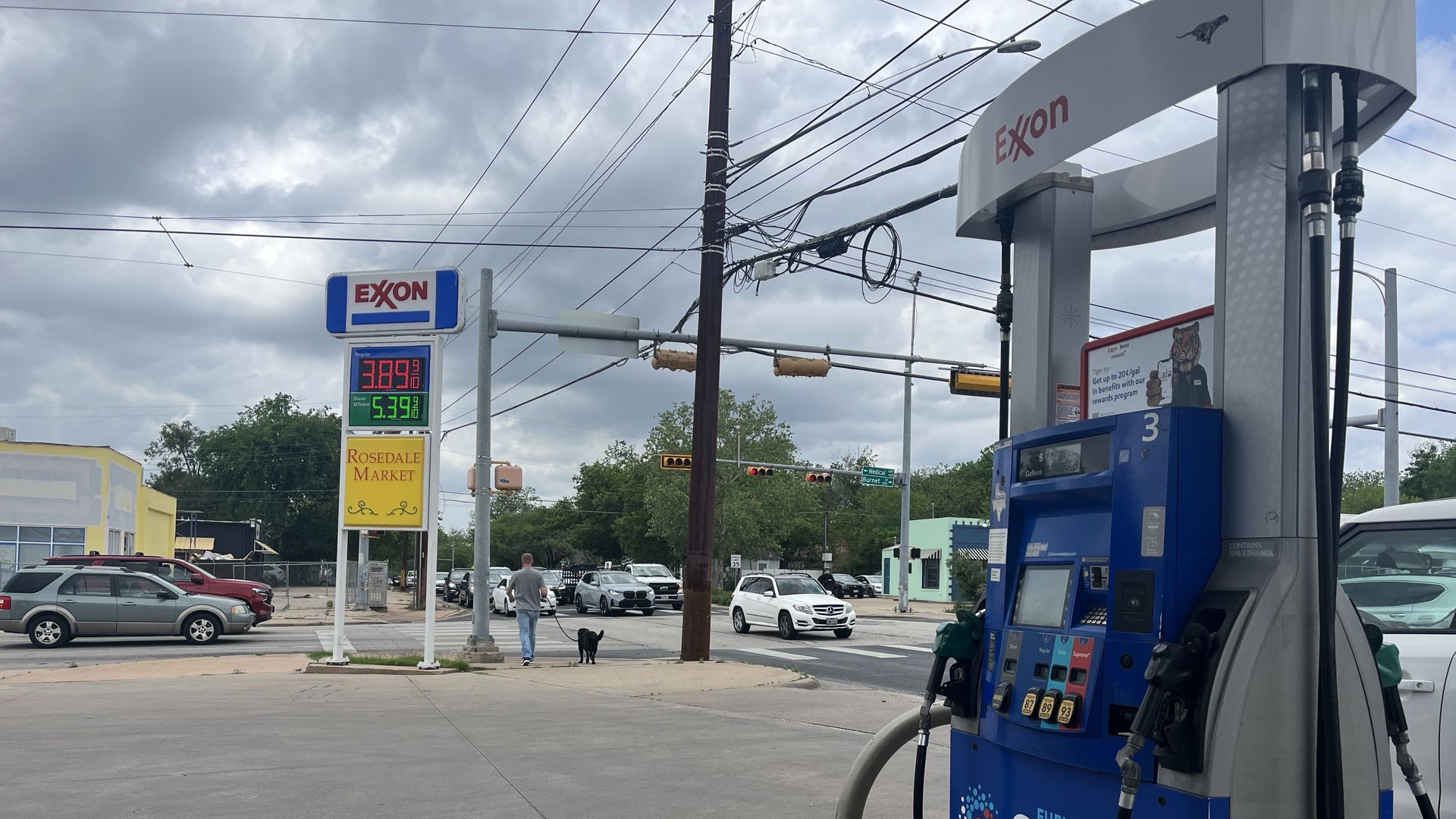 Exxon gas pump in the foreground on the right, blue Synergy branding and hoses. In the background, a man walks a dog near a yellow Rosedale Market sign, with cars and cloudy sky.