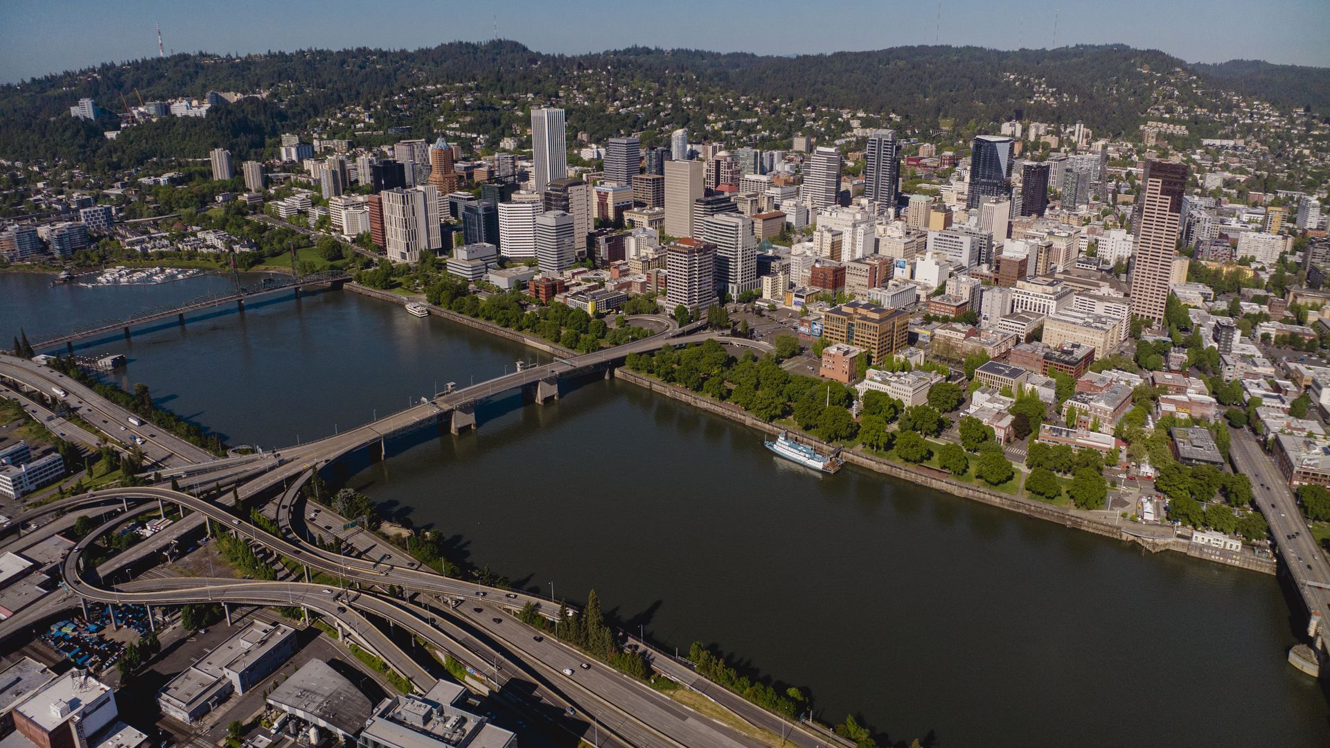 Aerial view of a riverfront city with many high-rise buildings, blue sky, two bridges over the water, complex highway interchanges, and forested hills in the distance.
