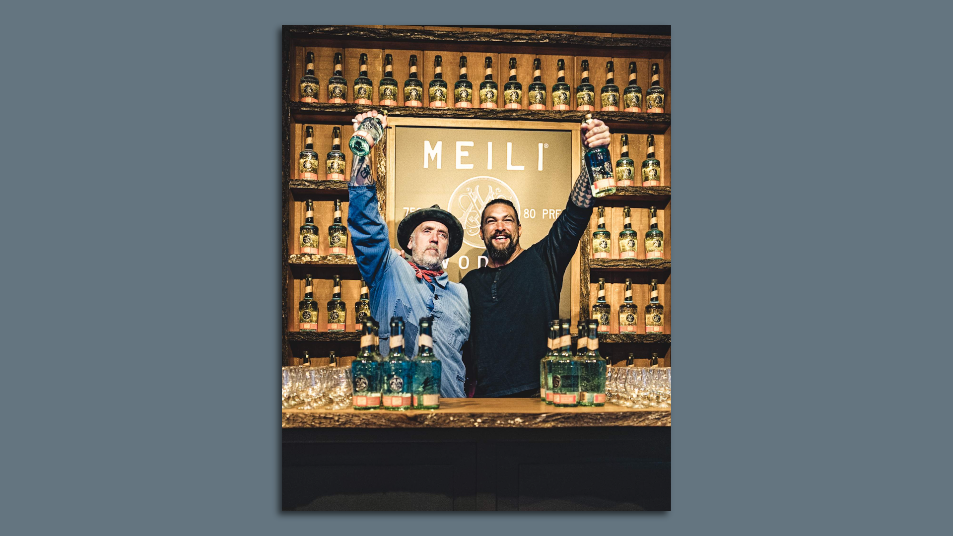 Two men embrace and hold up bottles of liquor in a celebratory pose 