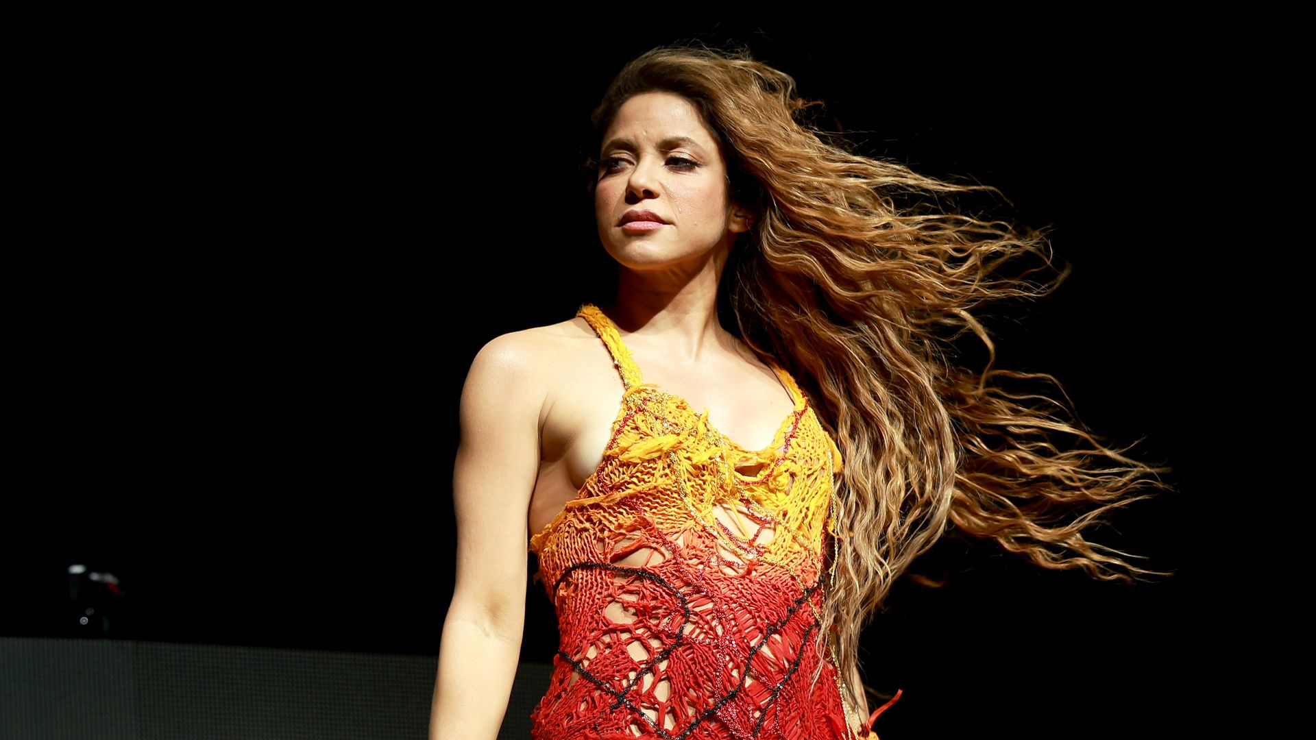 Female performer on stage with long, wavy hair blowing to the right, wearing a yellow-to-red crocheted dress, holding a microphone in her right hand against a dark background.