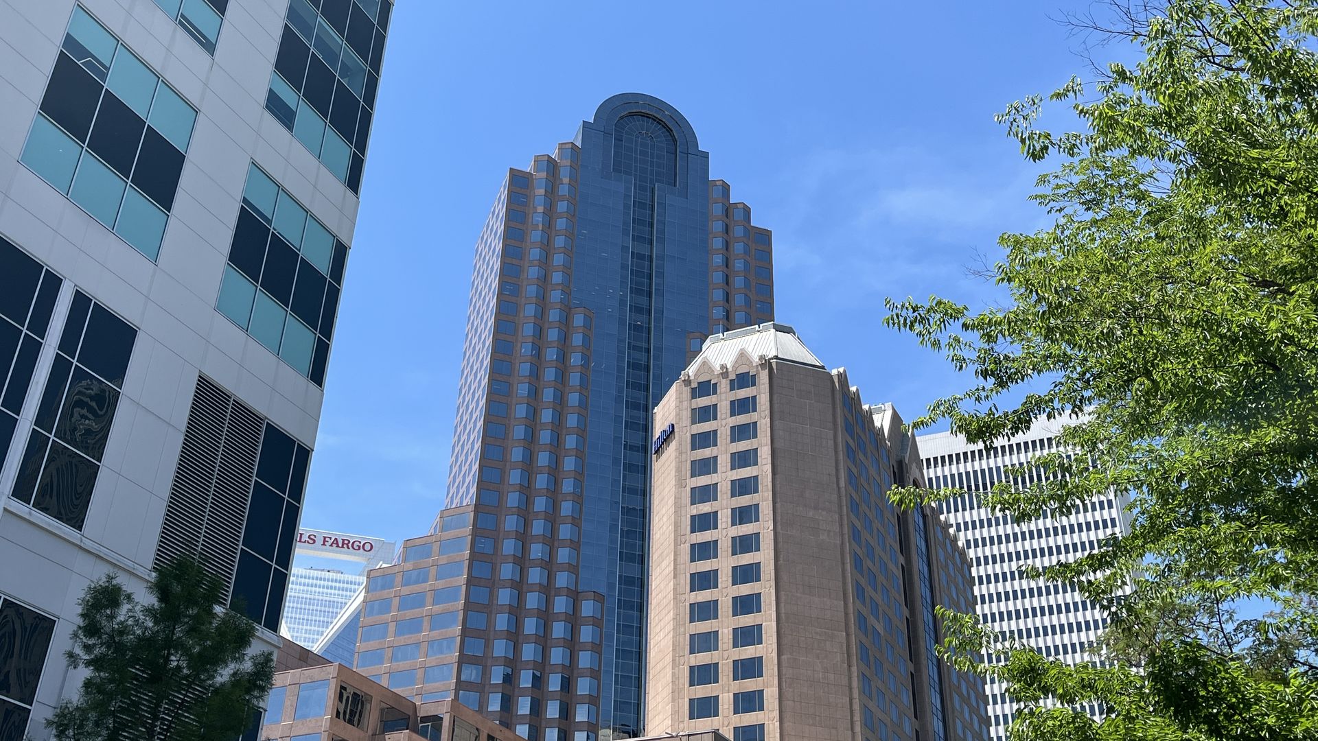 Tall glass office buildings rise toward a clear blue sky; a curved-top beige tower stands center, with green trees on the right and a partial Wells Fargo sign at the bottom left.