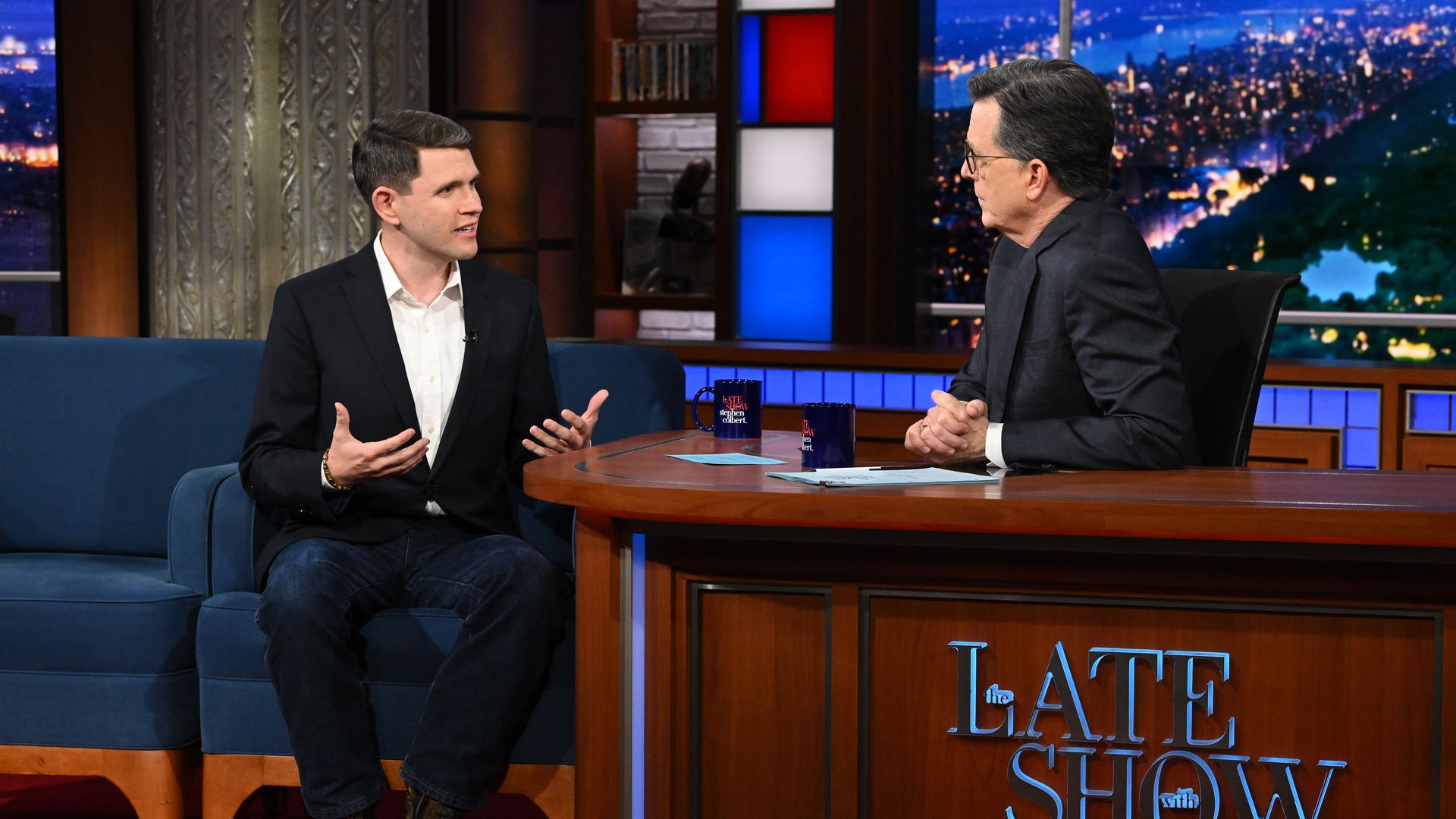 Talarico on the left talking with both of his hands raised, wearing a black suit jacket, white collared shirt and dark blue jeans while speaking to Colbert on the right wearing a black suit jacket 
