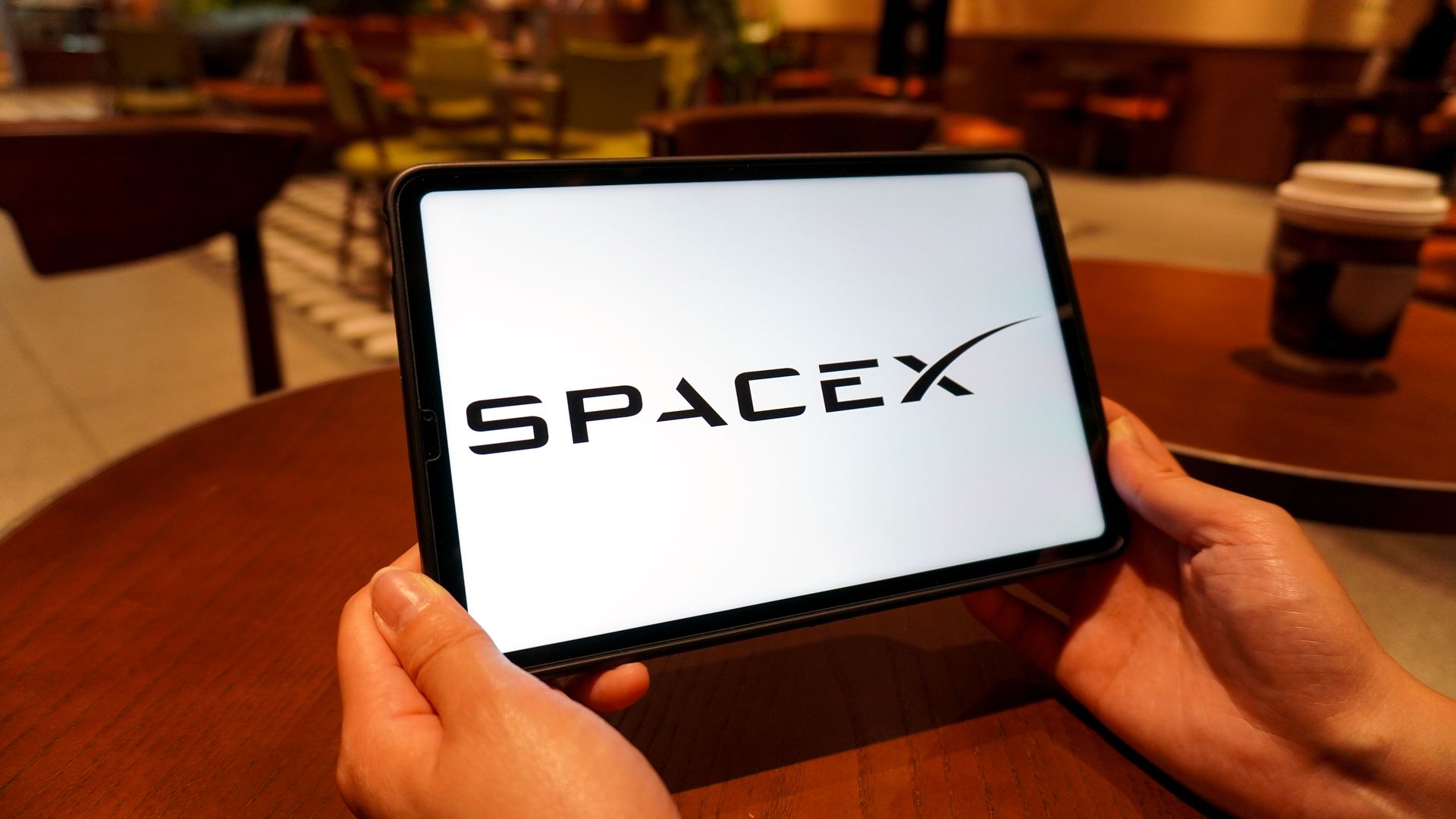 A close up of a smartphone screen displaying the SpaceX logo in bold black letters on white, held by two hands, with a blurred warm cafe interior in the background.