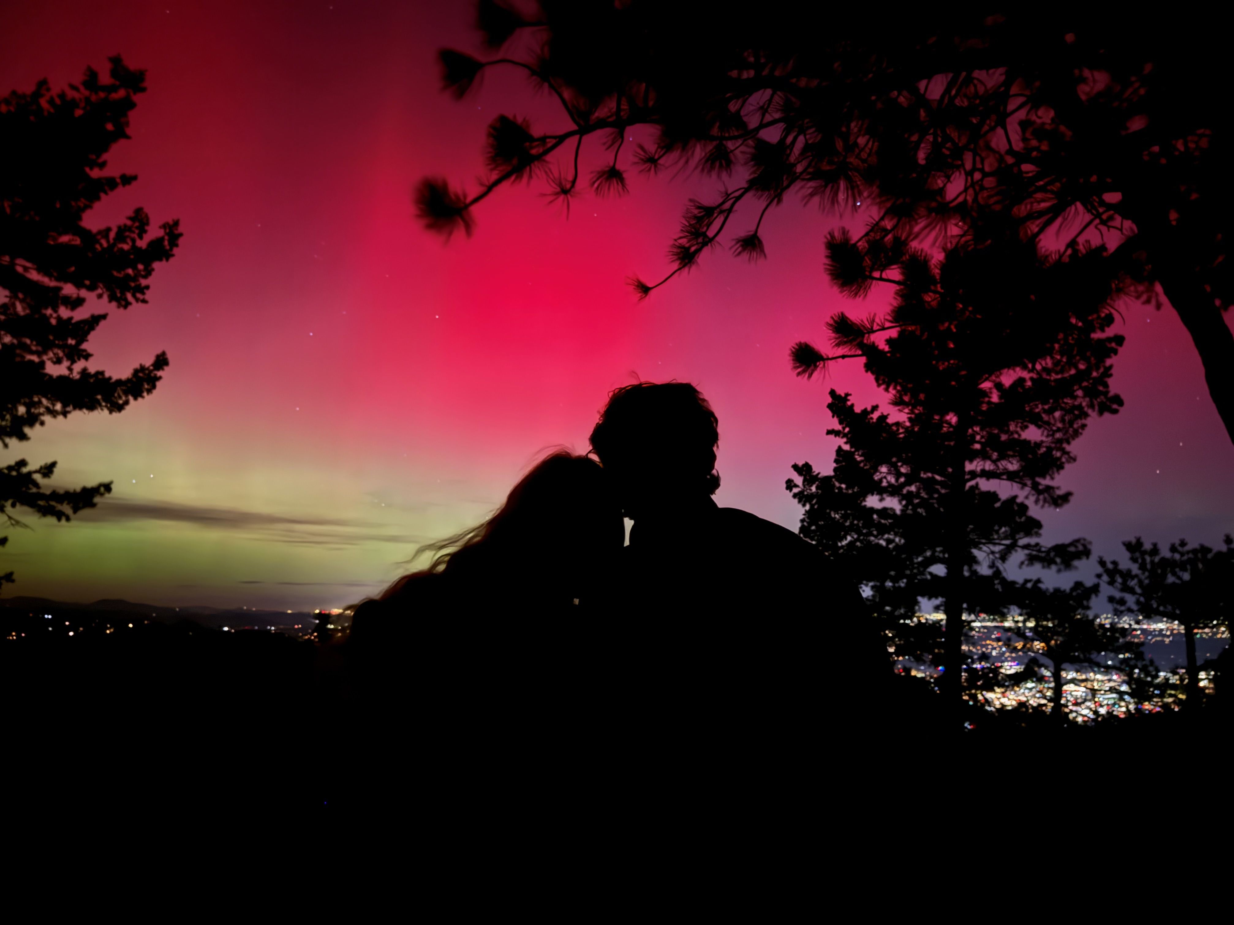 Silhouette of a couple leaning close against a backdrop of a vibrant pink and green aurora borealis above a cityscape, framed by dark tree branches at night.