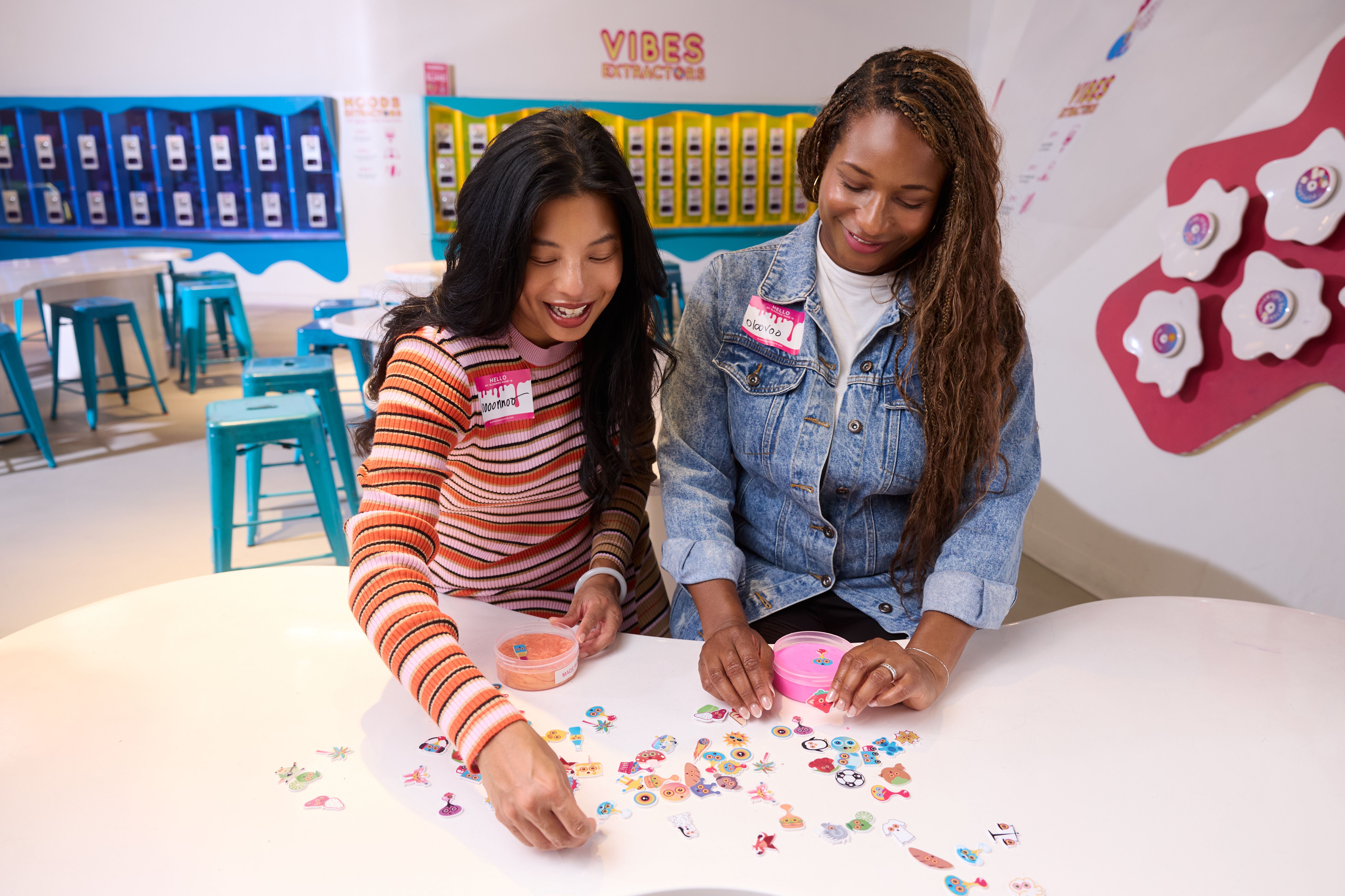 Two women sitting at a white table with colorful stickers, one wearing a striped orange-pink sweater and the other a denim jacket, in a brightly lit room with teal stools and wall displays.