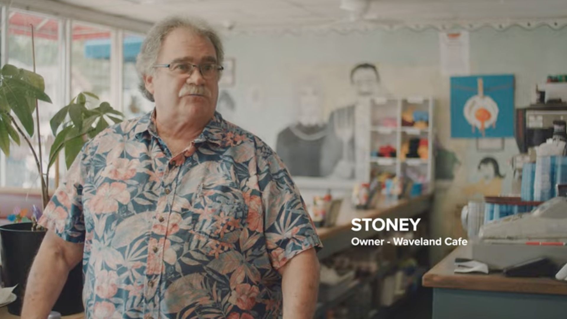 A gray-haired man with a mustache wearing a colorful floral Hawaiian shirt stands behind a cafe counter. A potted plant sits to his left, with shelves and posters in the background.