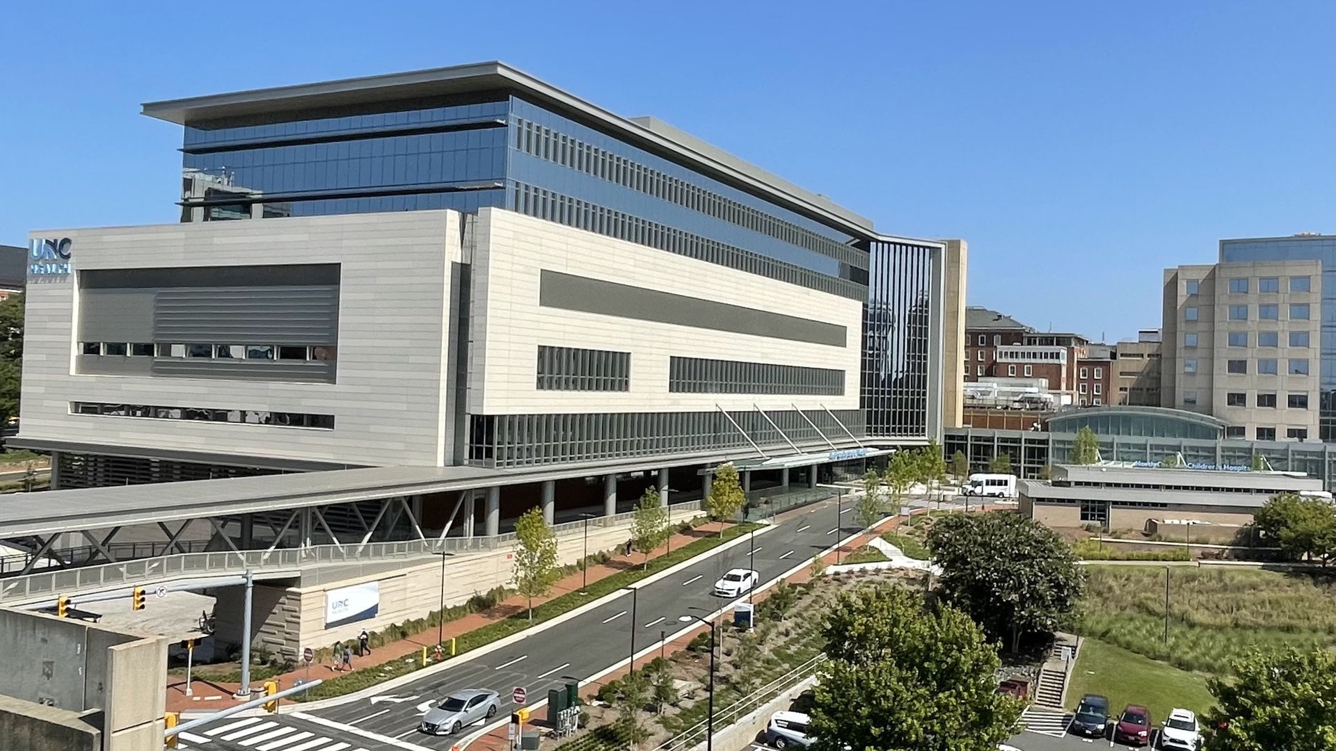 Modern multi-story hospital with glass and white paneling under clear blue sky, connected by an elevated walkway over a road with cars and surrounding green trees and parking.