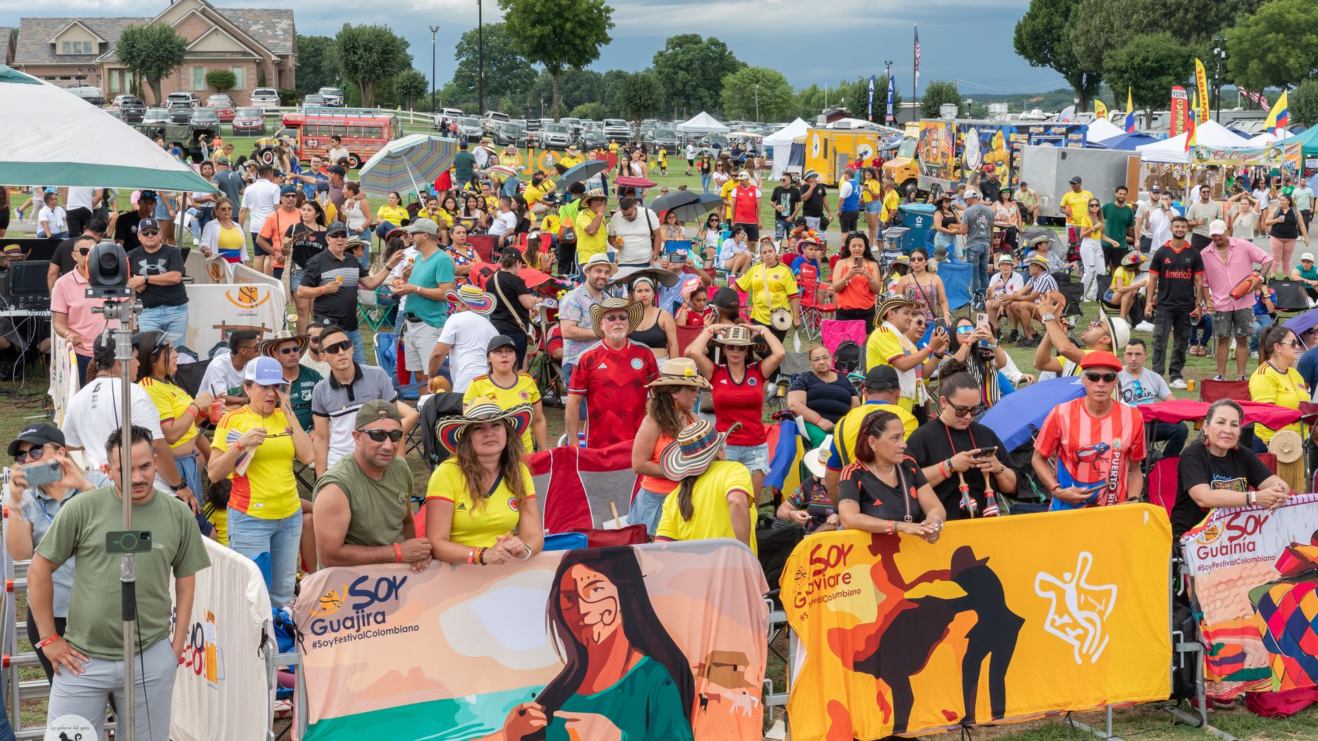 A large crowd of people enjoying an outdoor festival. Many attendees are wearing colorful attire, including yellow and red shirts, and traditional hats. They are gathered behind barriers with vibrant banners promoting "Soy Guajira" and "Soy Vivore" with festival names. Tents and stalls are visible i