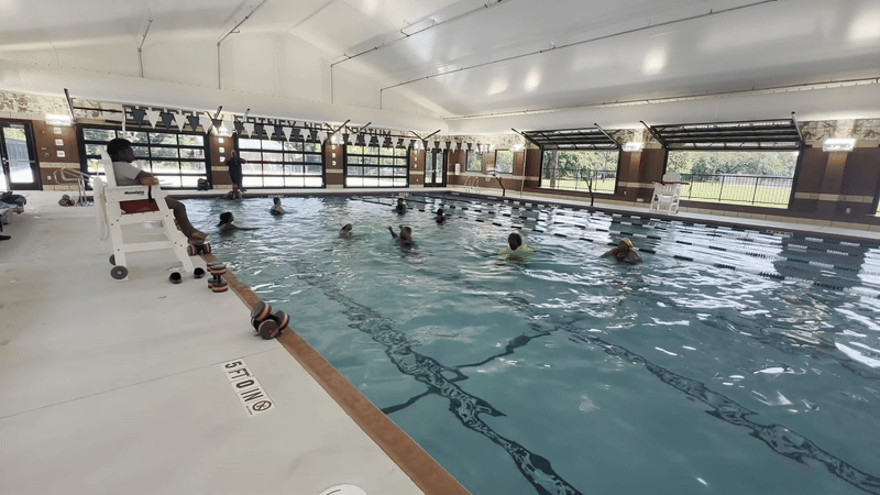 A group of about 8 women walk in a pool inside a natatorium with garage-style doors open.