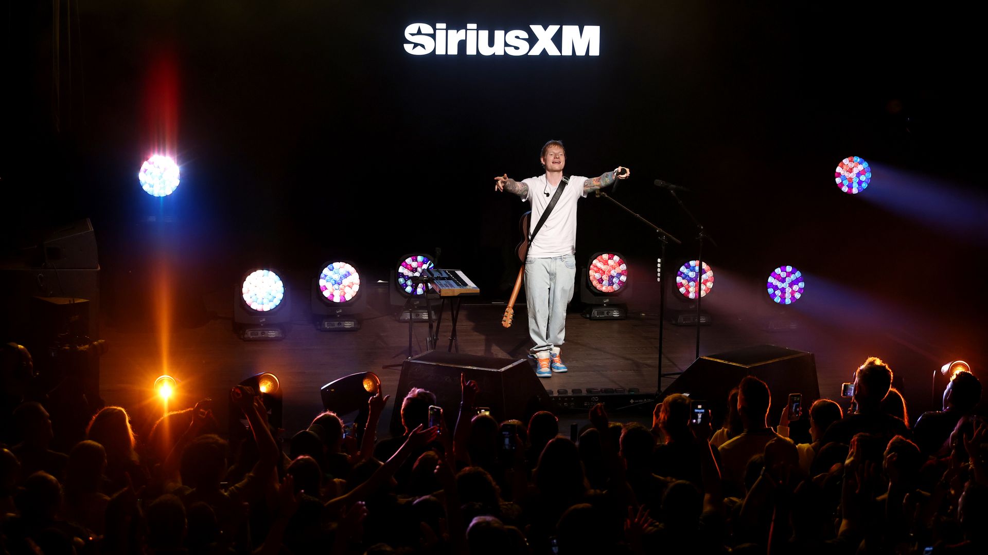 Singer on stage with guitar and microphone stands, arms outstretched, performing under bright spotlights