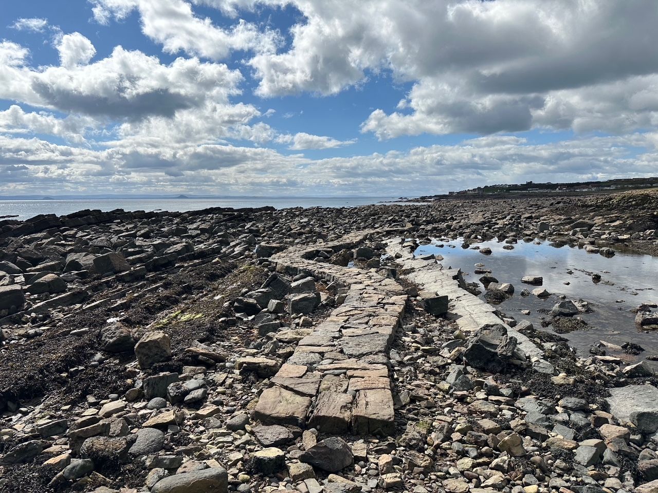 Rocky shoreline with a stone walkway leading toward the sea. Puffy clouds fill a blue sky; rocks and tidal pools dot the shore, with distant land on the horizon.