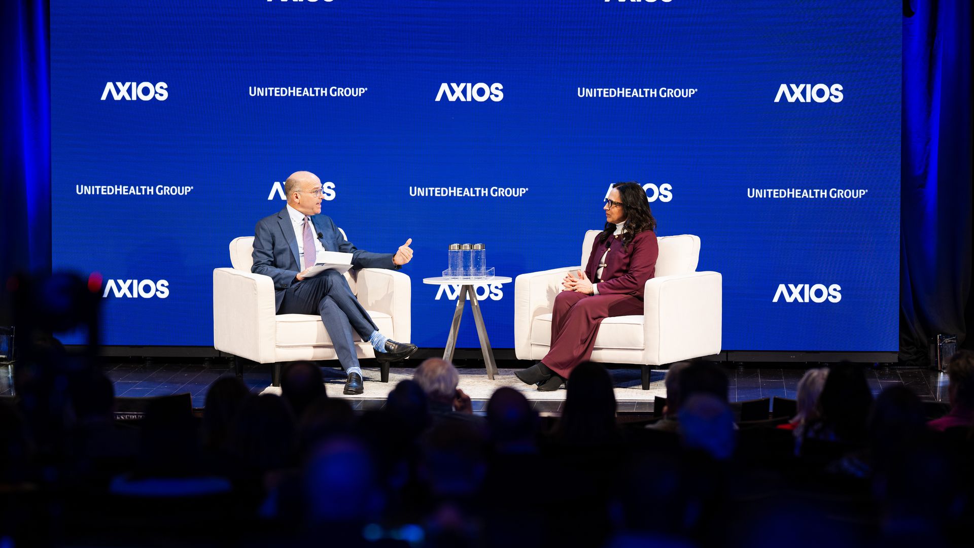 Two people seated on white armchairs on stage during an interview or discussion event with a blue backdrop featuring Axios and UnitedHealth Group logos and an audience in the foreground.