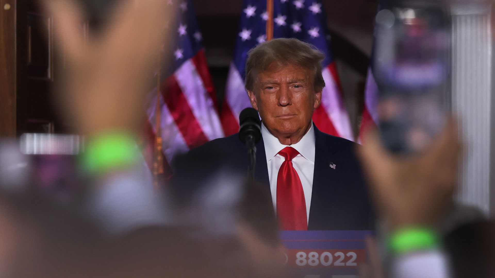 Donald Trump speaking at a lectern with spectators' hands raised in front of him. American flags are in the background.