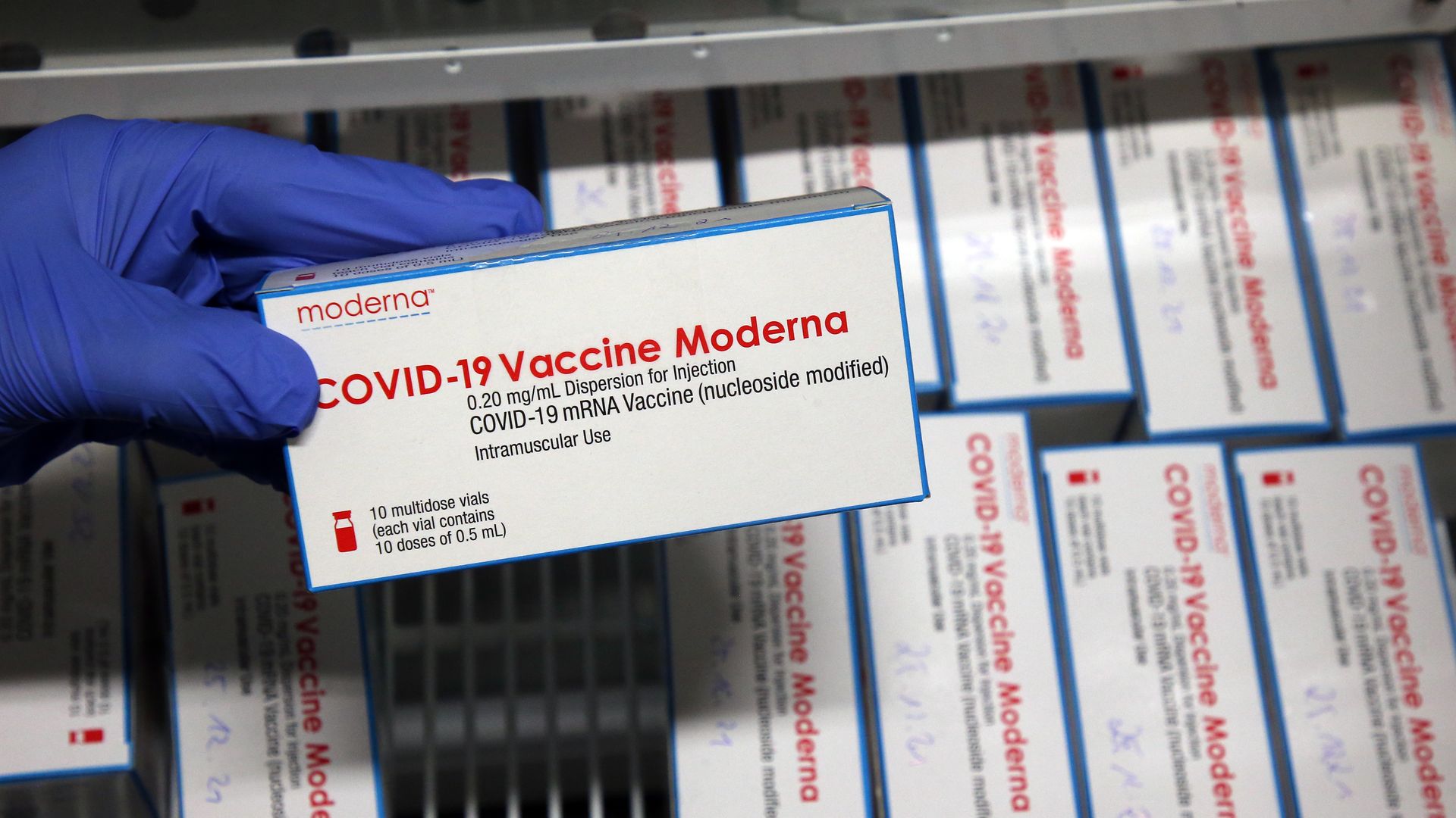 A hand holds a box of Moderna's COVID-19 vaccine, with other boxes in the background.