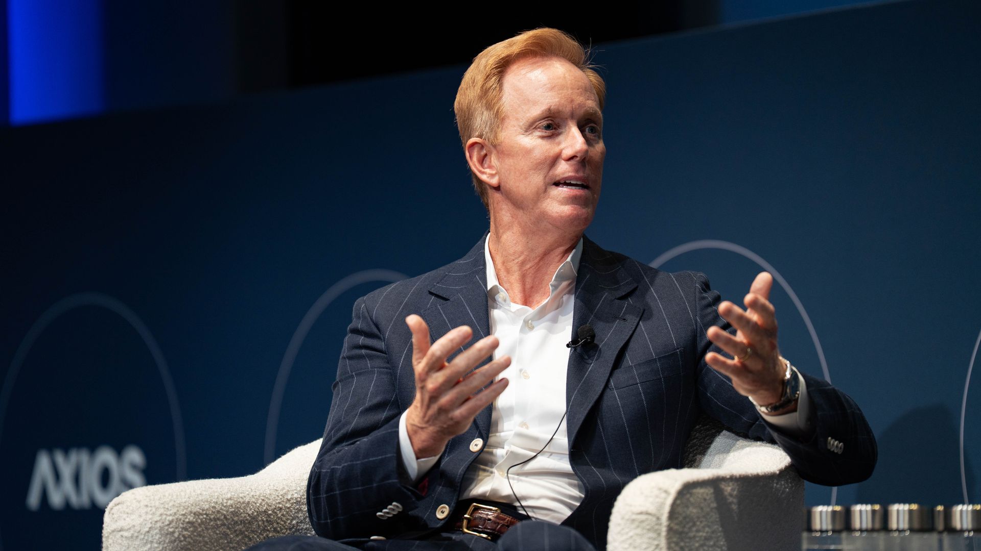 Middle-aged man with light brown hair wearing a dark pinstripe suit and white shirt, speaking and gesturing with hands while seated on a white armchair at an event with blue background.