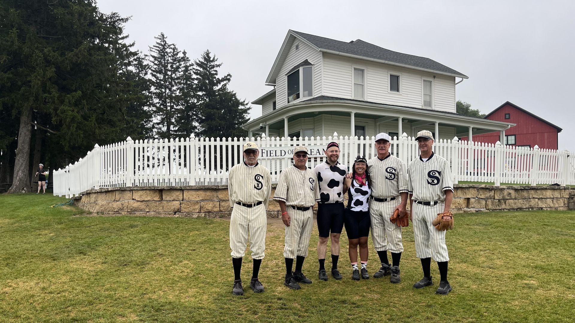 Six people in cream pinstripe baseball uniforms pose before a white picket fence, a two-story house, and a red barn on a grassy field; a sign reads "FIELD OF DREAMS".