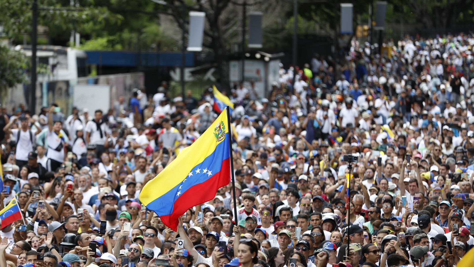 Hundreds of people march very closely together in Caracas, Venezuela. Someone holds up a large Venezuelan flag