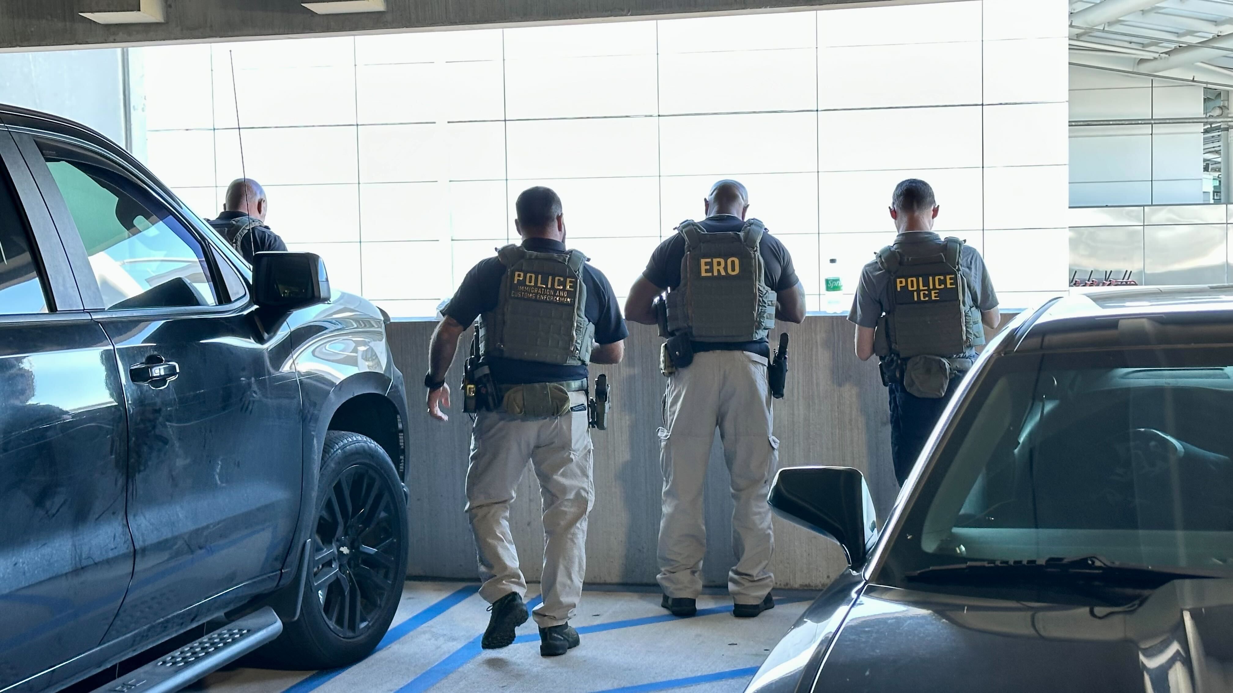 Three officers in tactical vests labeled POLICE ICE and ERO stand with their backs to the camera in a parking garage, flanked by parked cars and a concrete barrier against a bright white wall.
