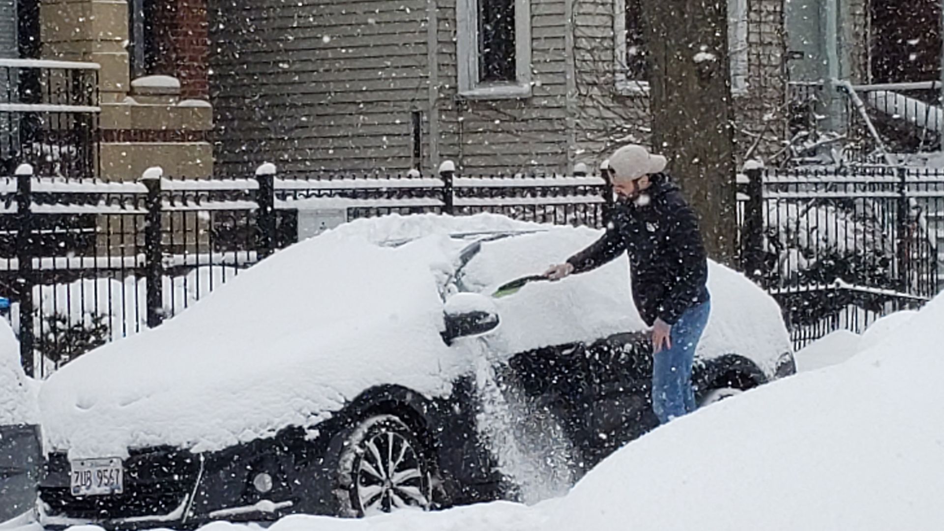 Person in black jacket, blue jeans, and beige cap brushing snow off a black car covered in thick snow on a snowy street with houses and fences in the background during snowfall.
