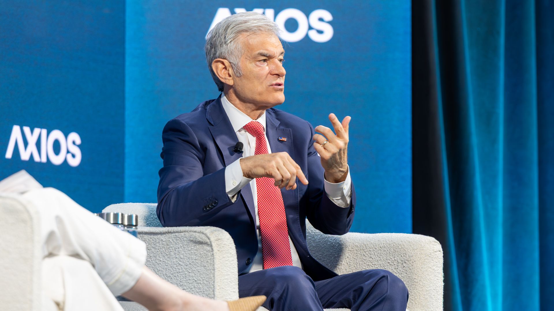 A grey-haired man in a navy suit and red tie sits on a gray chair on a blue stage with the Axios branding in the background, gesturing with his hands during a discussion.