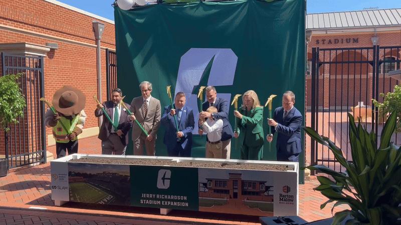 Seven people wearing formal and business attire hold gold-colored shovels, participating in a groundbreaking ceremony for Jerry Richardson Stadium Expansion with green backdrop and brick buildings.
