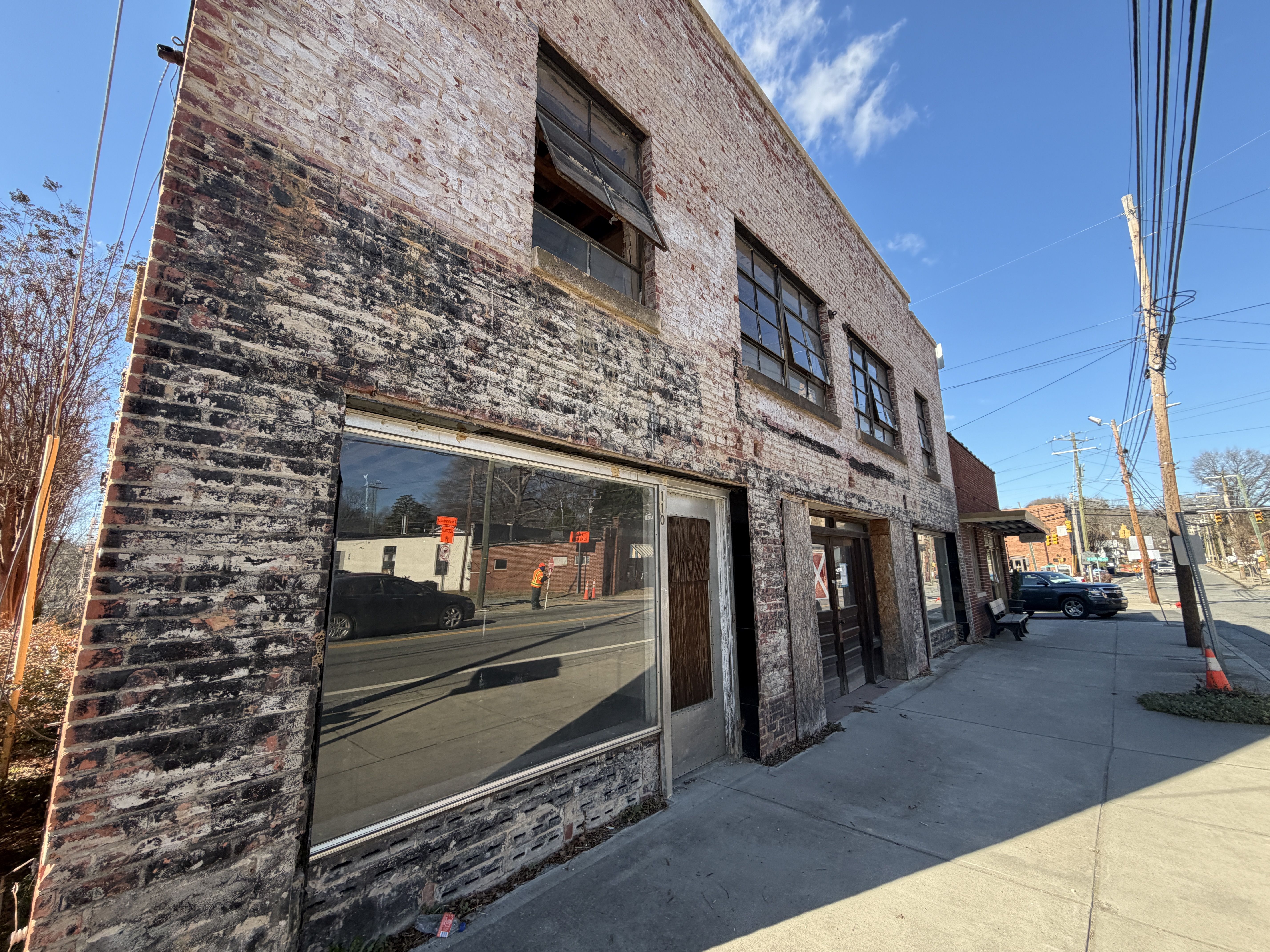 Weathered, two-story brick building with faded white and black paint, broken windows, and boarded doors on a sunny street with clear blue sky and parked cars.
