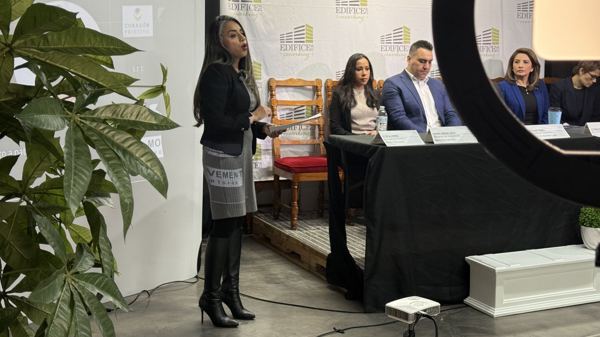 A woman speaks to an audience while surrounded by a screen and a plant. To her right, four people are seated. 