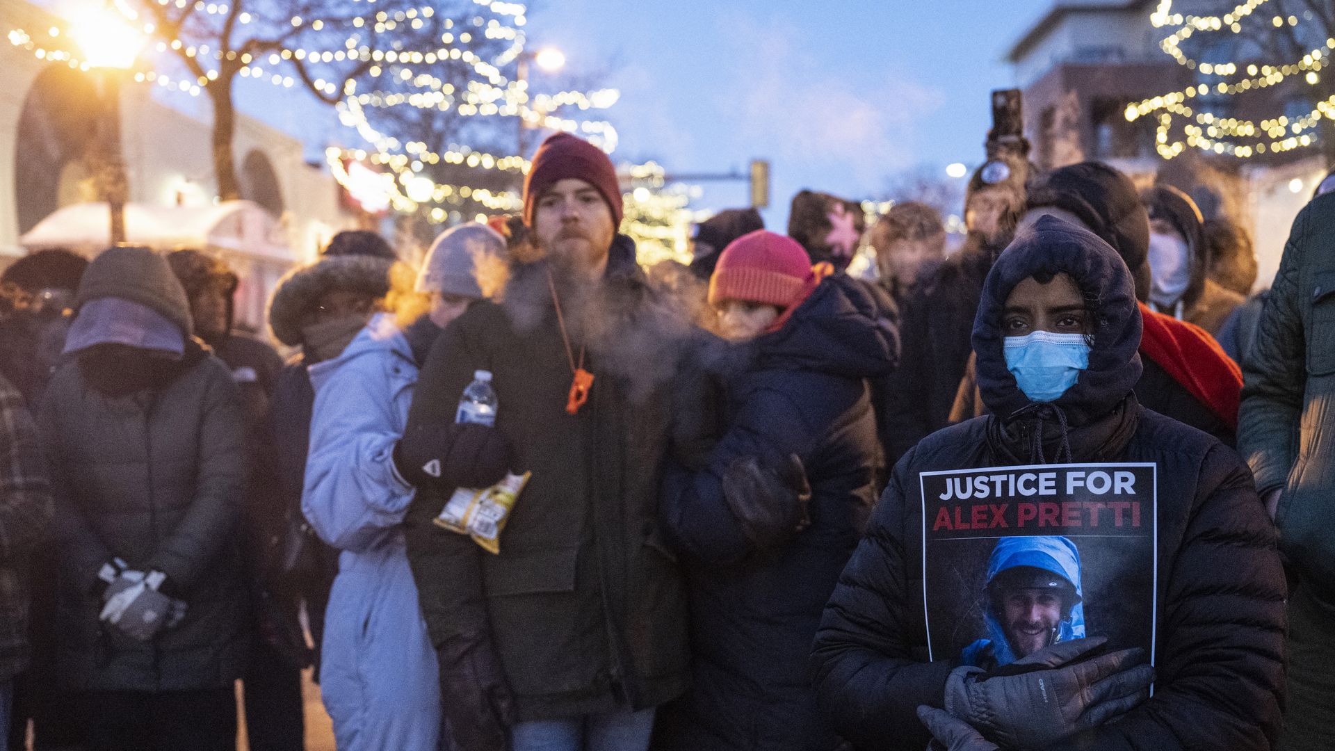 A group of people stand together at a makeshift memorial for a man shot by federal agents in Minnesota. One person, wearing a blue mask, holds a sign that says "JUSTICE FOR ALEX PRETTI."