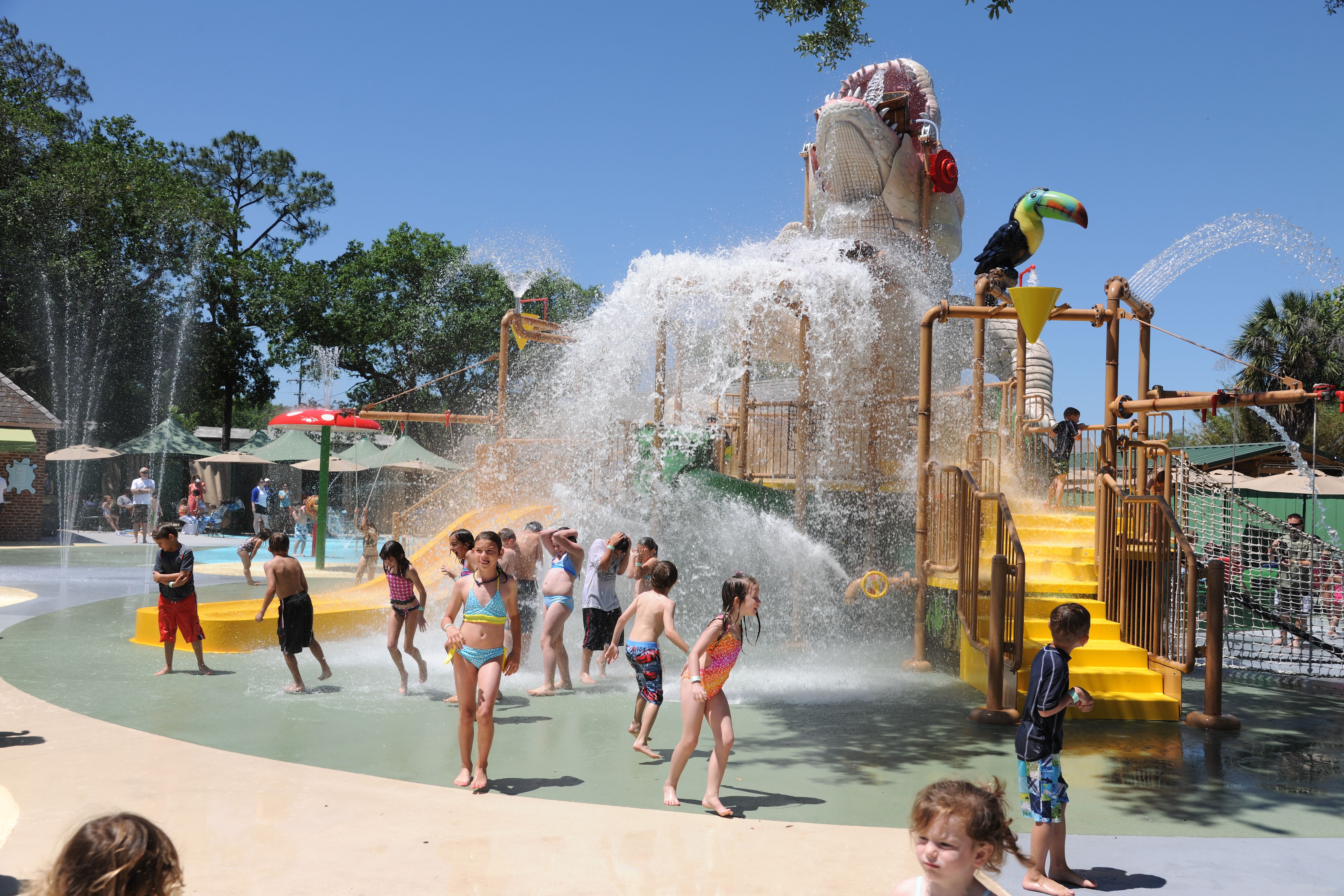 Photo shows the Cool Zoo on a sunny day. The white alligator's head is pointed up and water dumps beneath it as children in bathing suits play.
