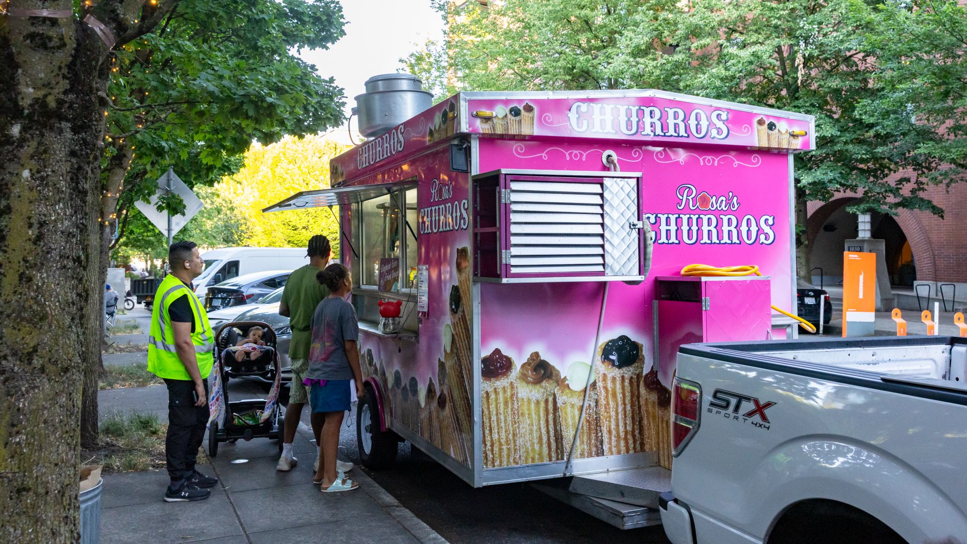 Pink food truck named Rosa's Churros with images of churros, people standing in line, and a baby in a stroller on a city sidewalk surrounded by trees and buildings.