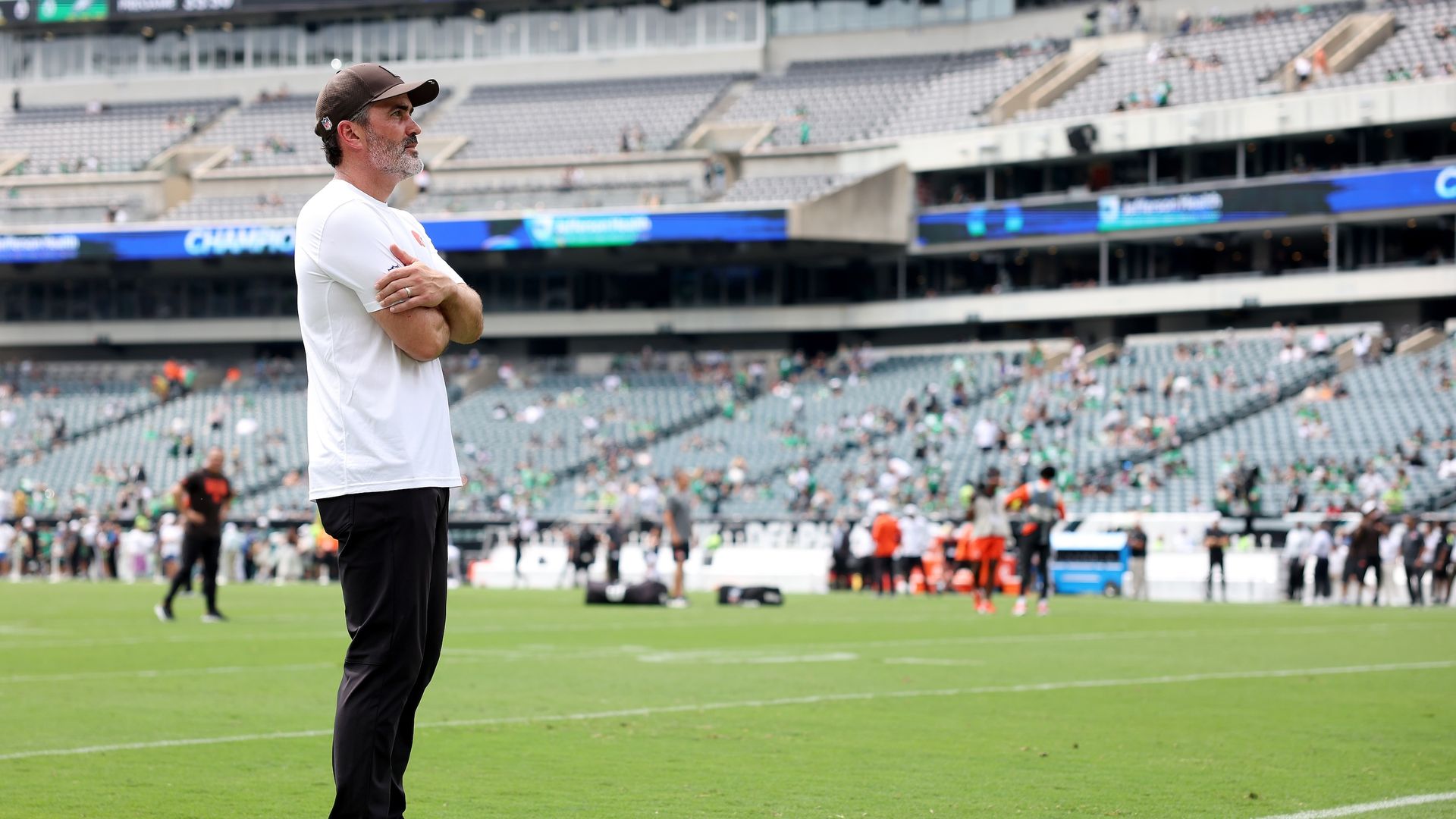 Man in white t-shirt, black pants, and black-and-white sneakers stands with arms crossed on a football field with blurred players and empty stadium seats in the background.