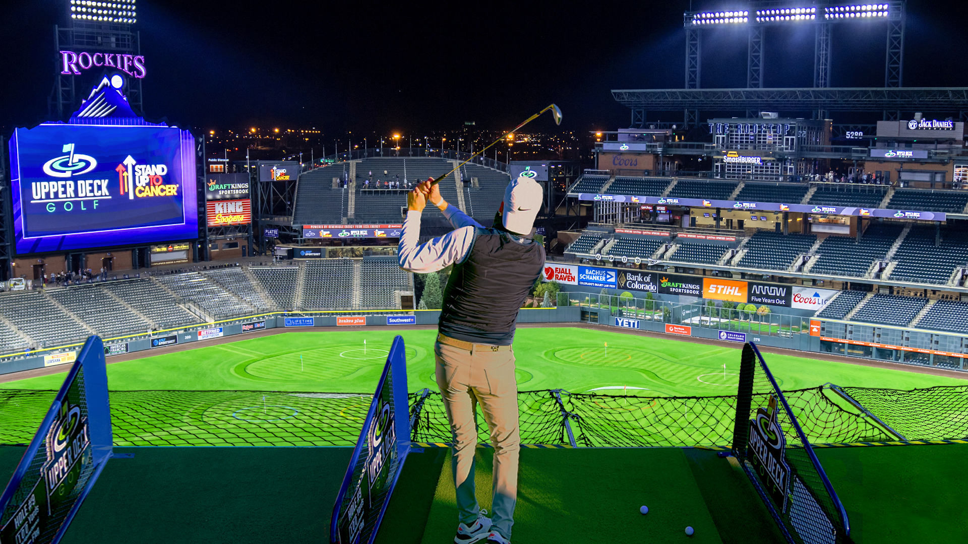 A man swings a golf club while overlooking a baseball stadium. 
