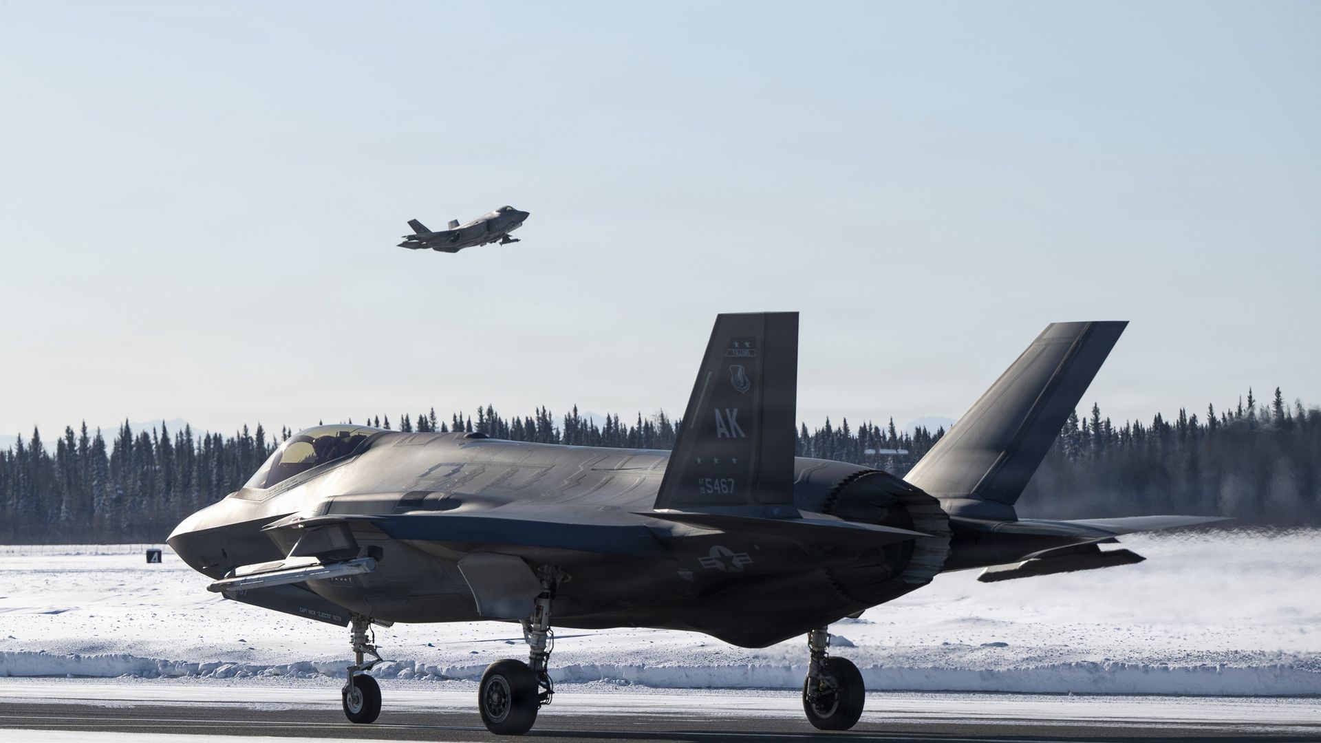 Two gray fighter jets at a snowy airbase with pine trees and mountains in the background. One is on the runway and one is taking off into clear blue sky.