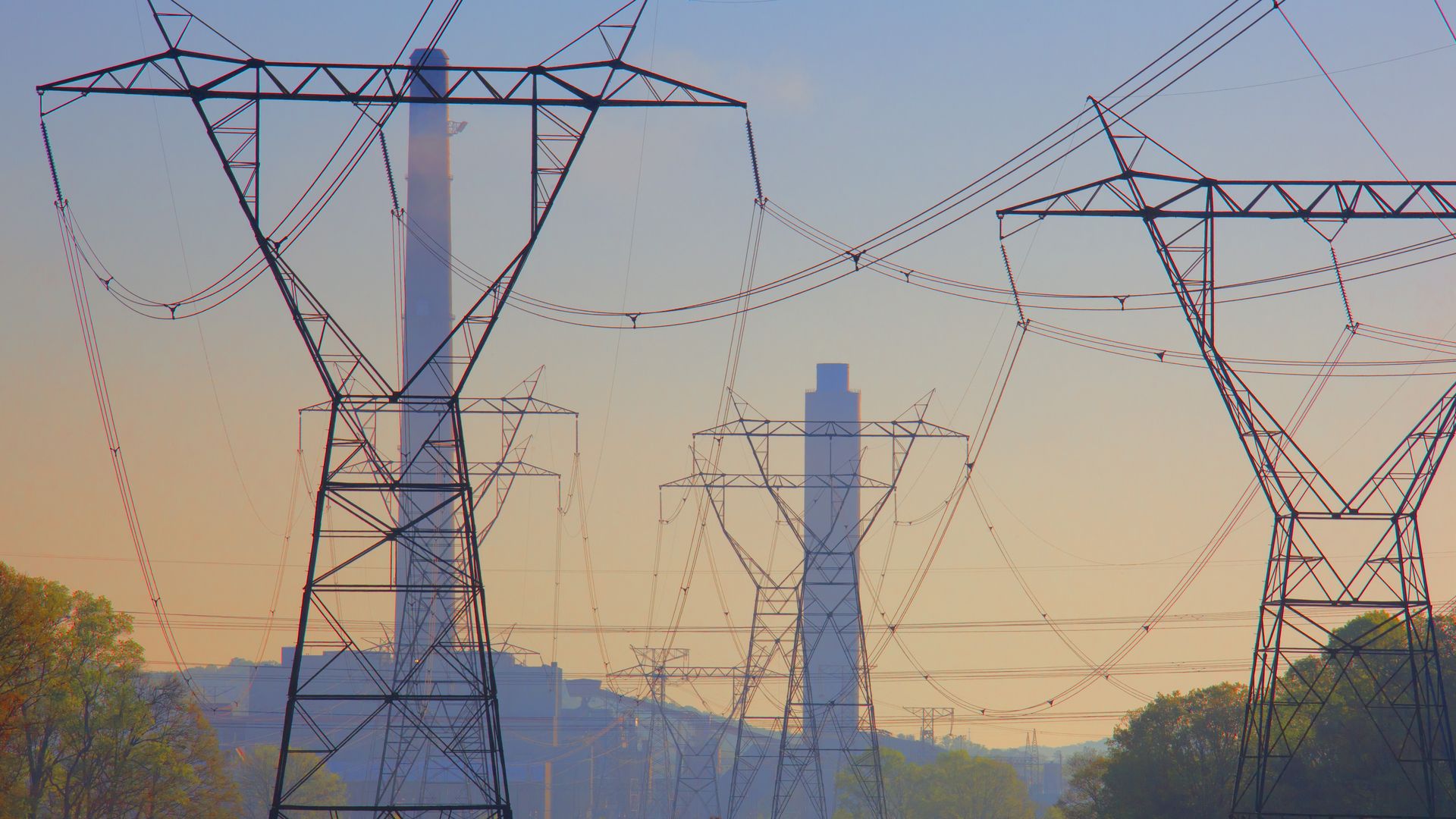 A photo of tall transmission lines, set against smokestacks for the retired Bull Run Fossil plant, against a sunset sky.