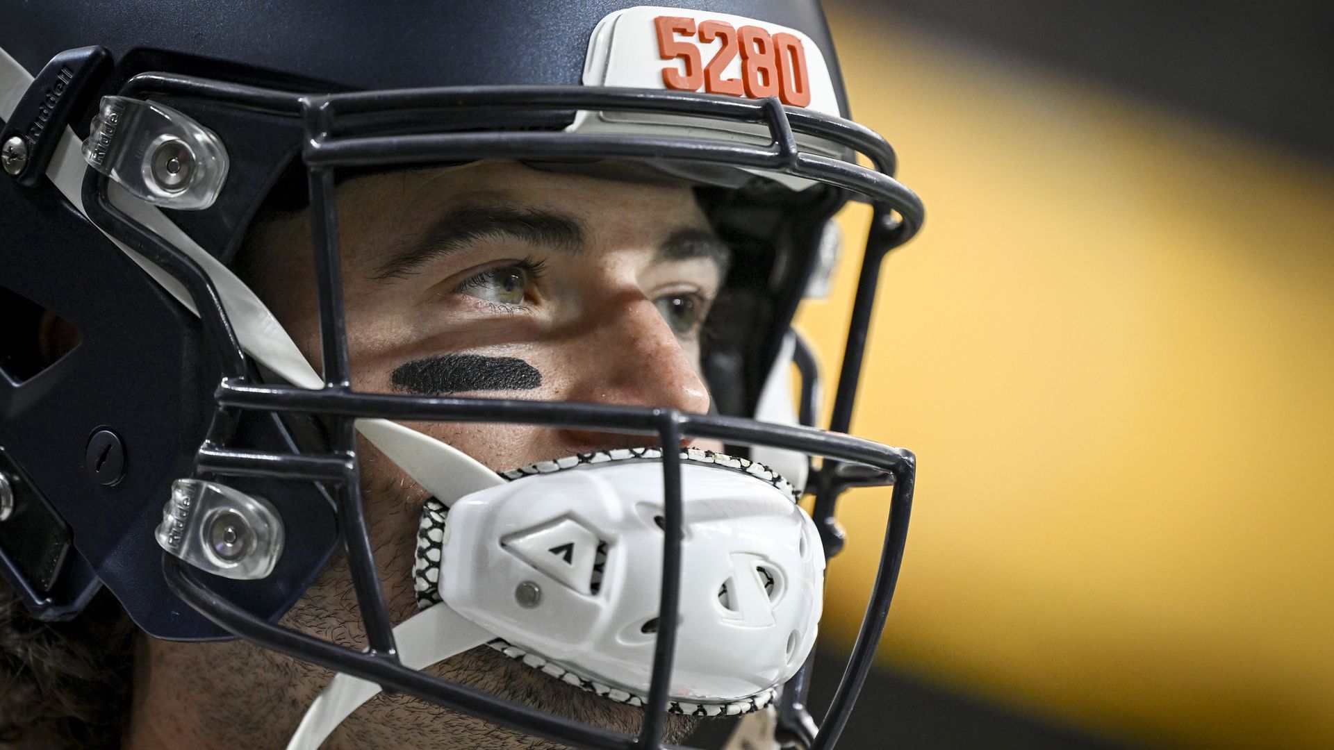 Close-up of a football player wearing a navy helmet with orange number 5280 and black eye paint, looking focused with a white mouthguard attached to the face mask.
