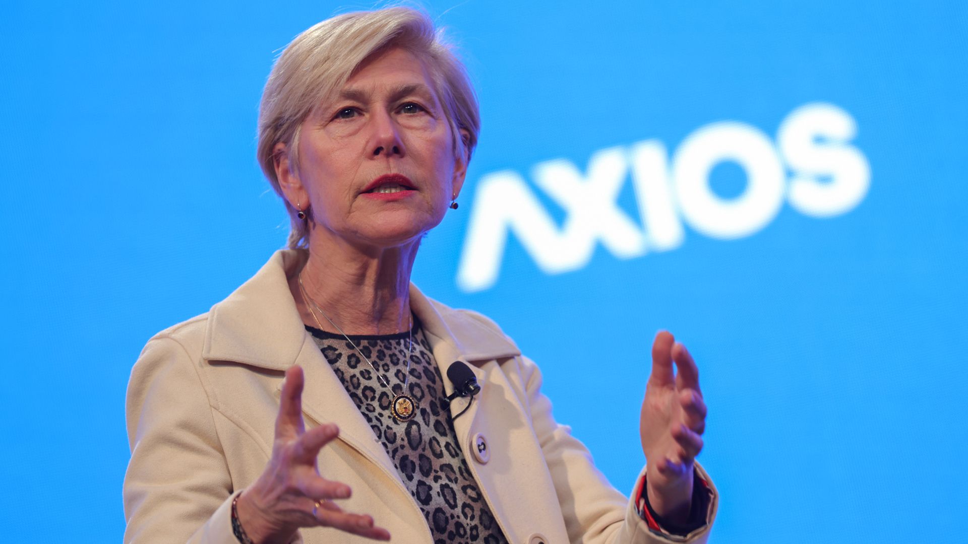 Middle-aged woman with short blonde hair wearing a cream blazer over a leopard-print blouse speaks on stage, gesturing with her hands, microphone clipped, against a blue backdrop featuring the Axios logo.