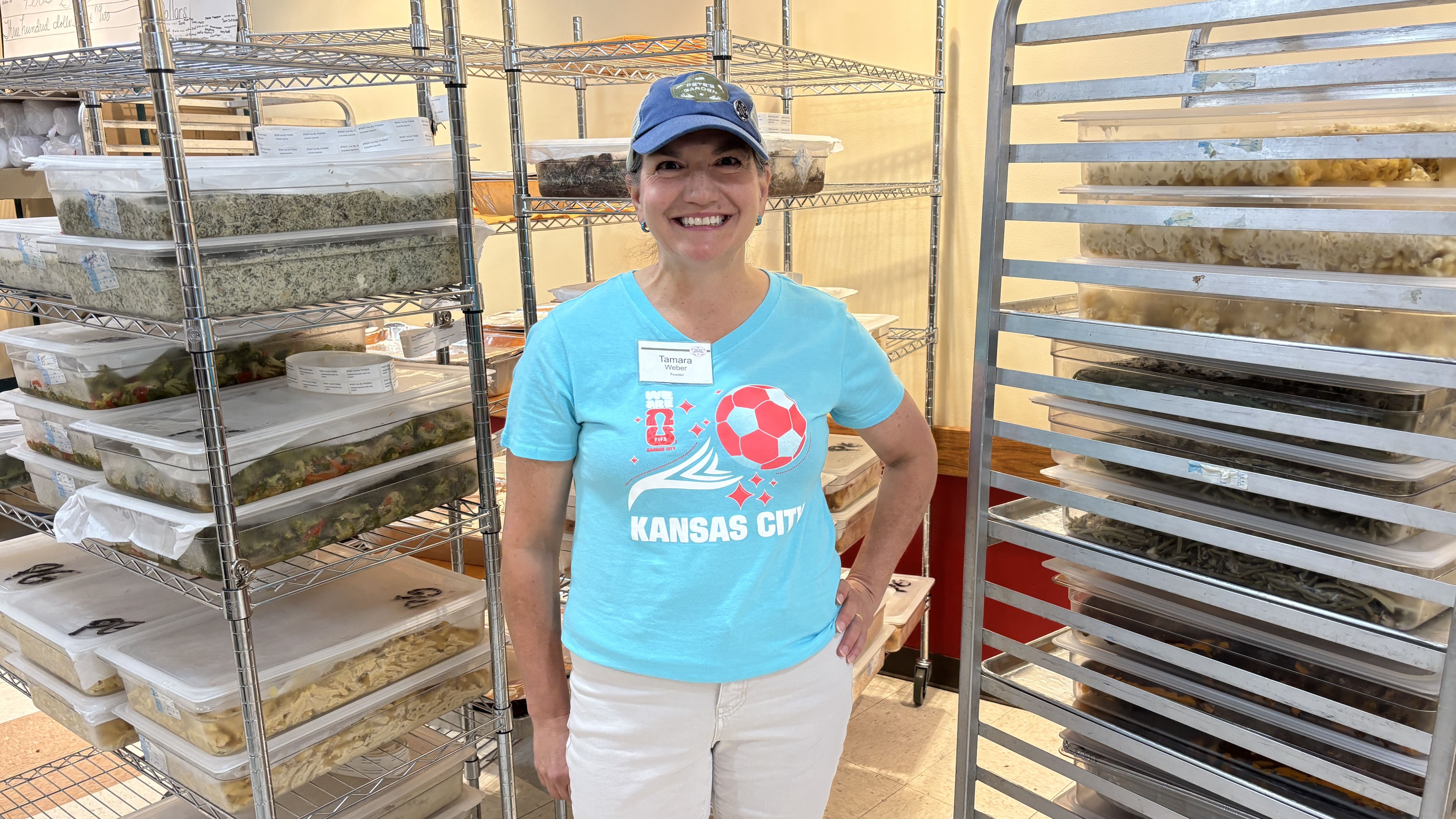 Tamara Weber smiles in a light blue Kansas City shirt and cap and stands in a kitchen storage area with metal shelves and clear containers of salads, grains, and pasta trays.