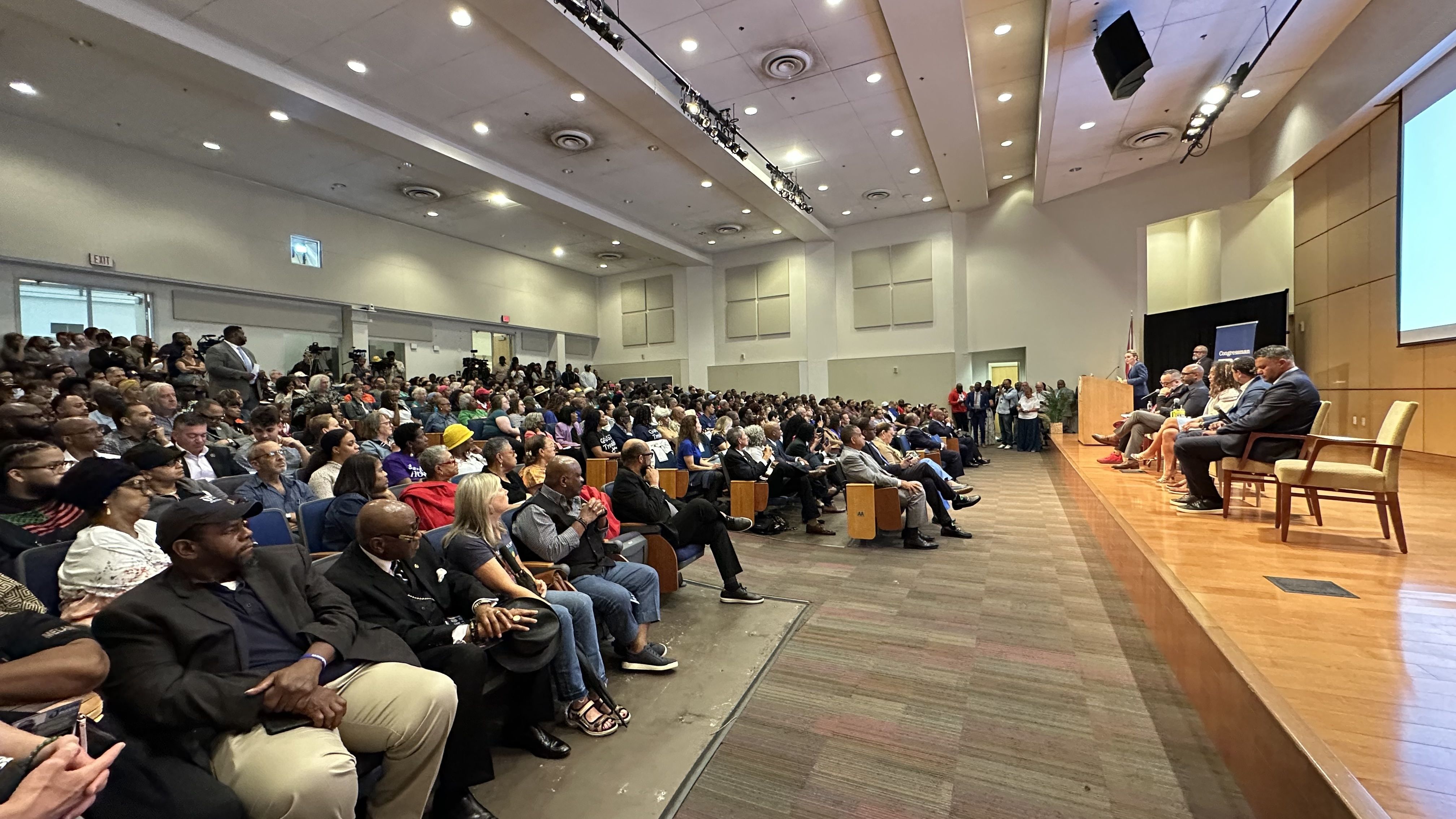 A large conference hall with a panel of speakers on a wooden stage to the right and a crowded audience seated in rows on the left.